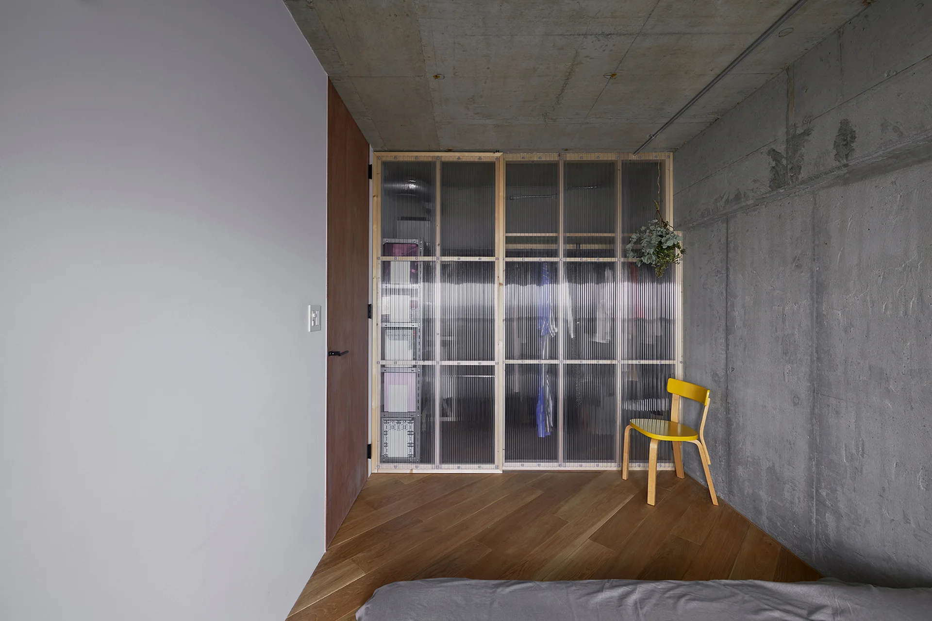 A minimalist bedroom features raw concrete walls and ceiling, angled hardwood flooring, a yellow wooden chair, and a translucent fluted glass closet.
