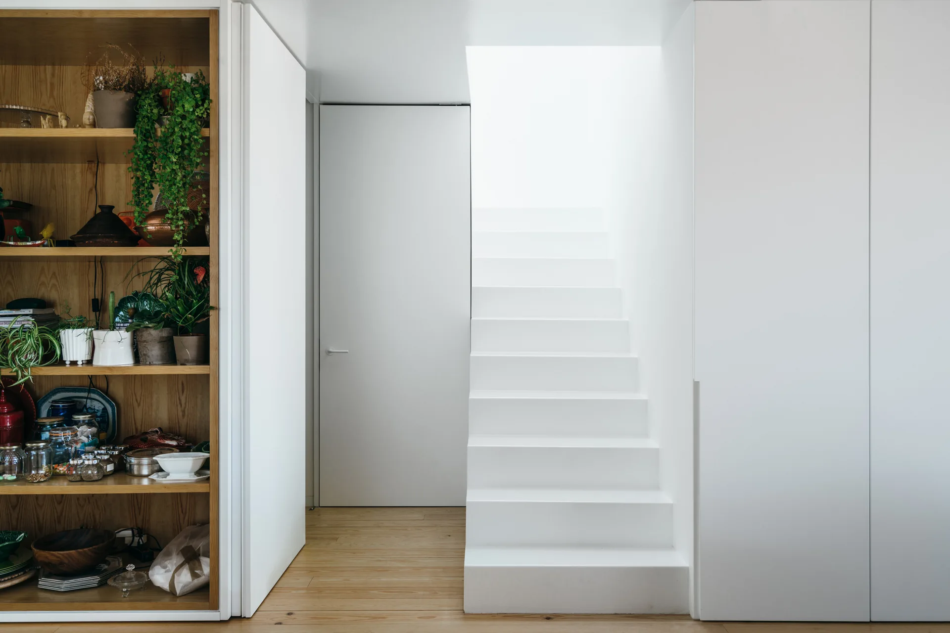 A modern white staircase is positioned between a tall light-wood shelving unit filled with potted plants and seamless white wall panels.
