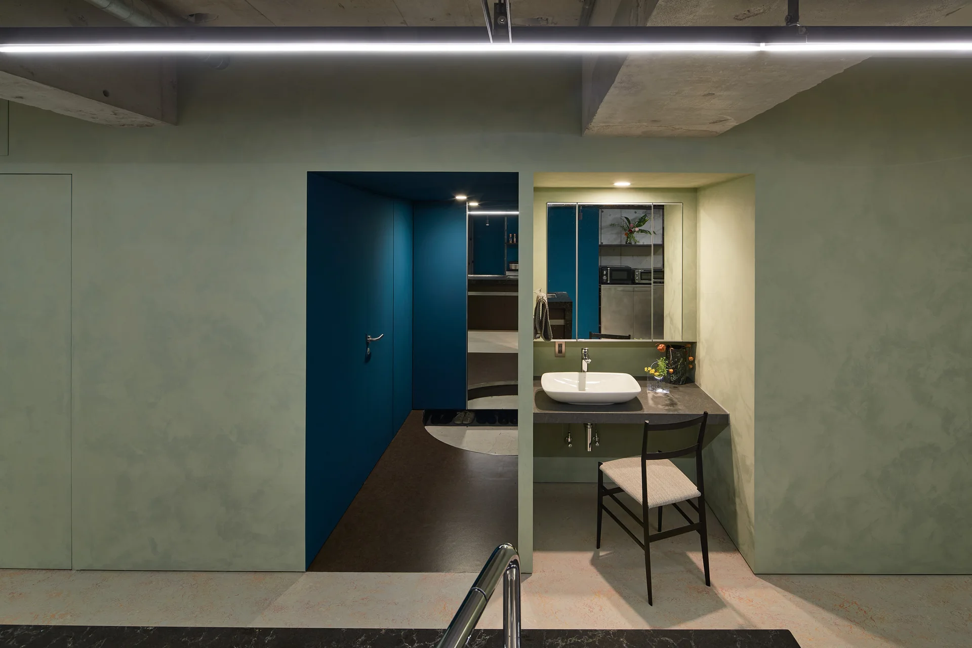 A bathroom vanity featuring a white vessel sink is recessed into a sage green plaster wall next to a deep blue doorway under an exposed concrete ceiling.