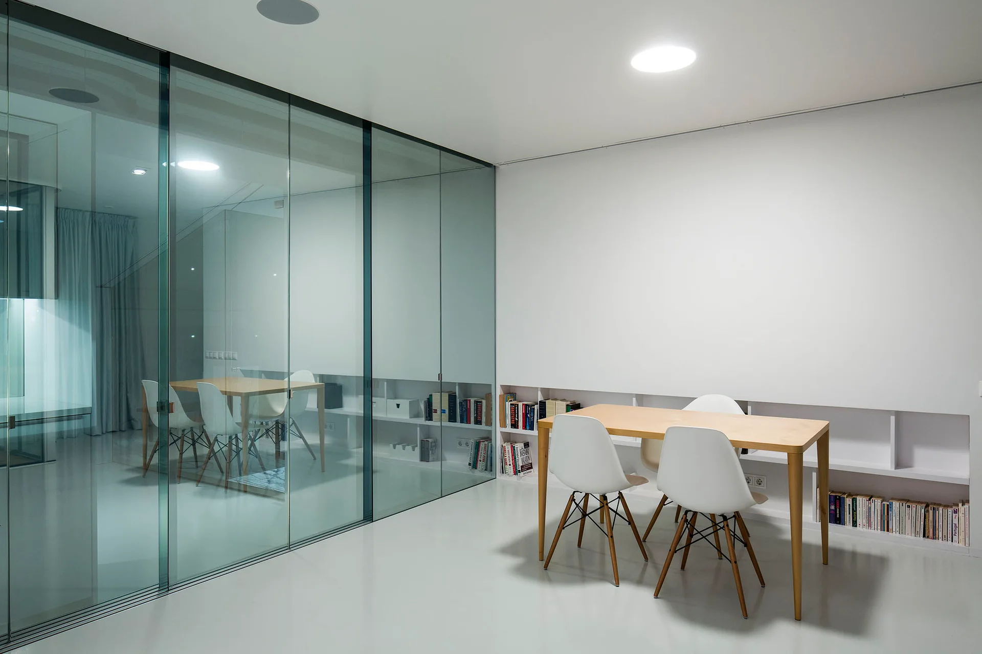 A minimalist dining room features a light wood table, white molded chairs, a low recessed bookshelf, and a floor-to-ceiling glass partition wall.