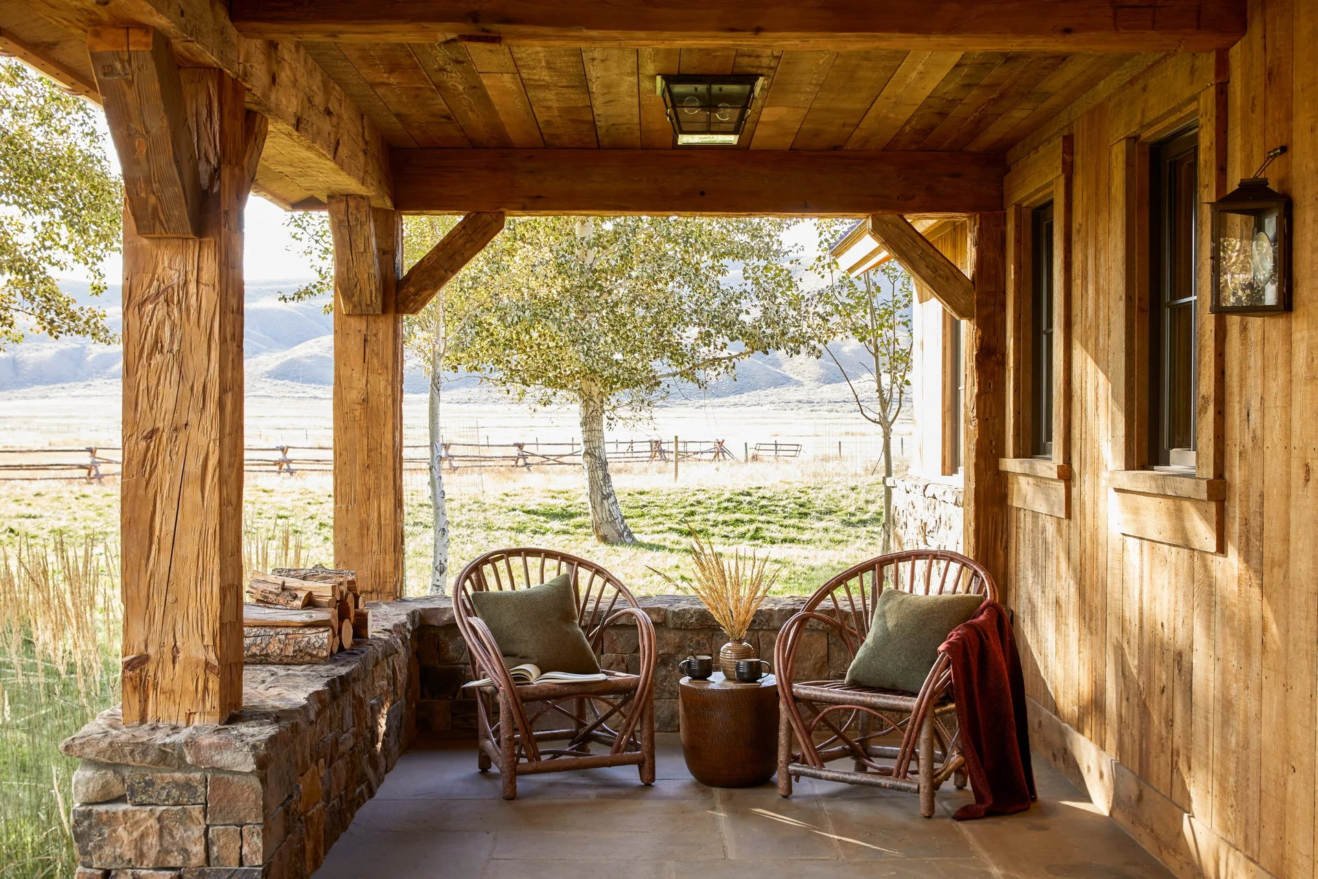 A rustic covered porch features two twig armchairs with moss-green pillows, a stone half-wall, and thick timber posts looking out at a field.