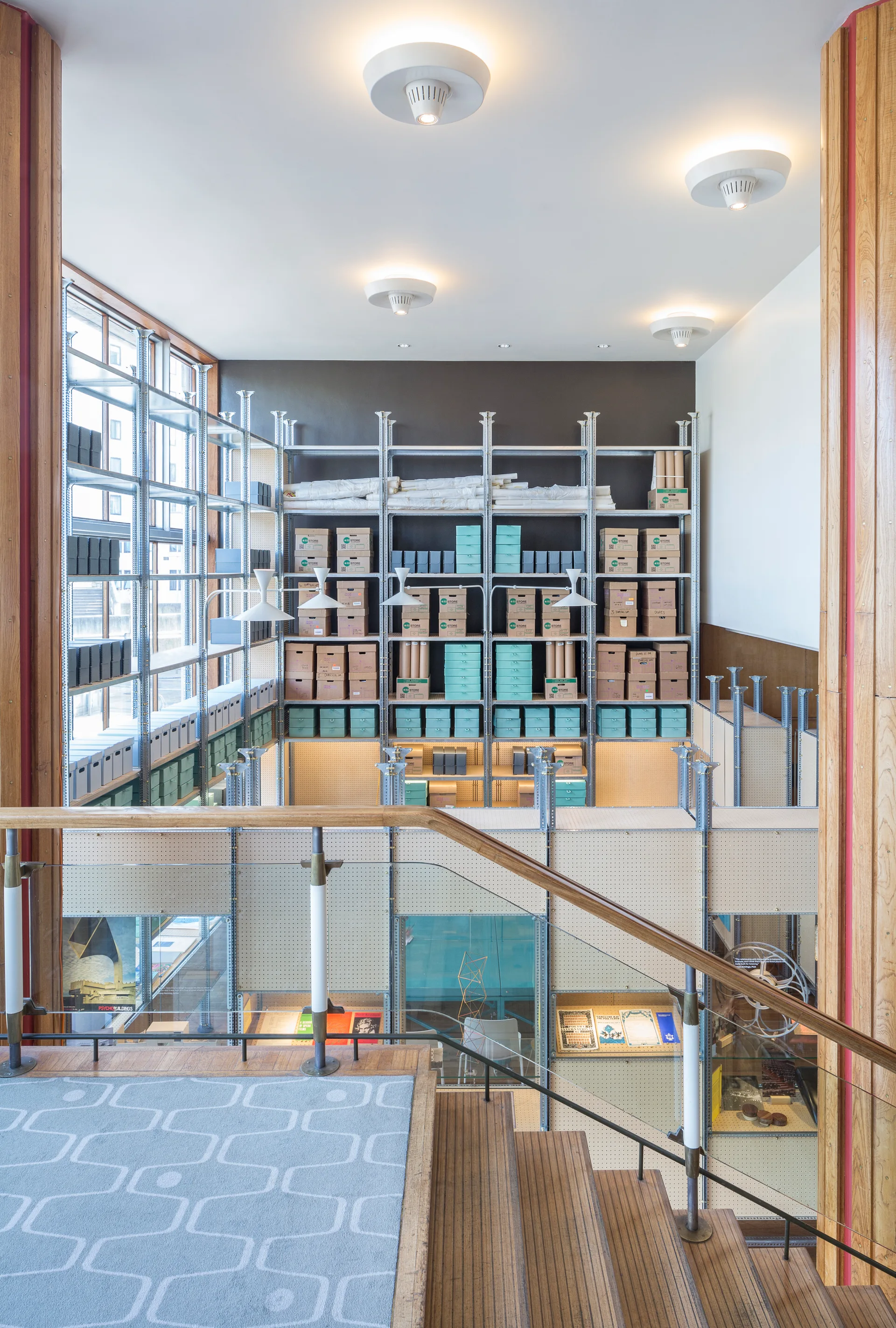 A view from a wooden staircase landing with a patterned blue rug, looking down toward large industrial metal shelving units filled with storage boxes.