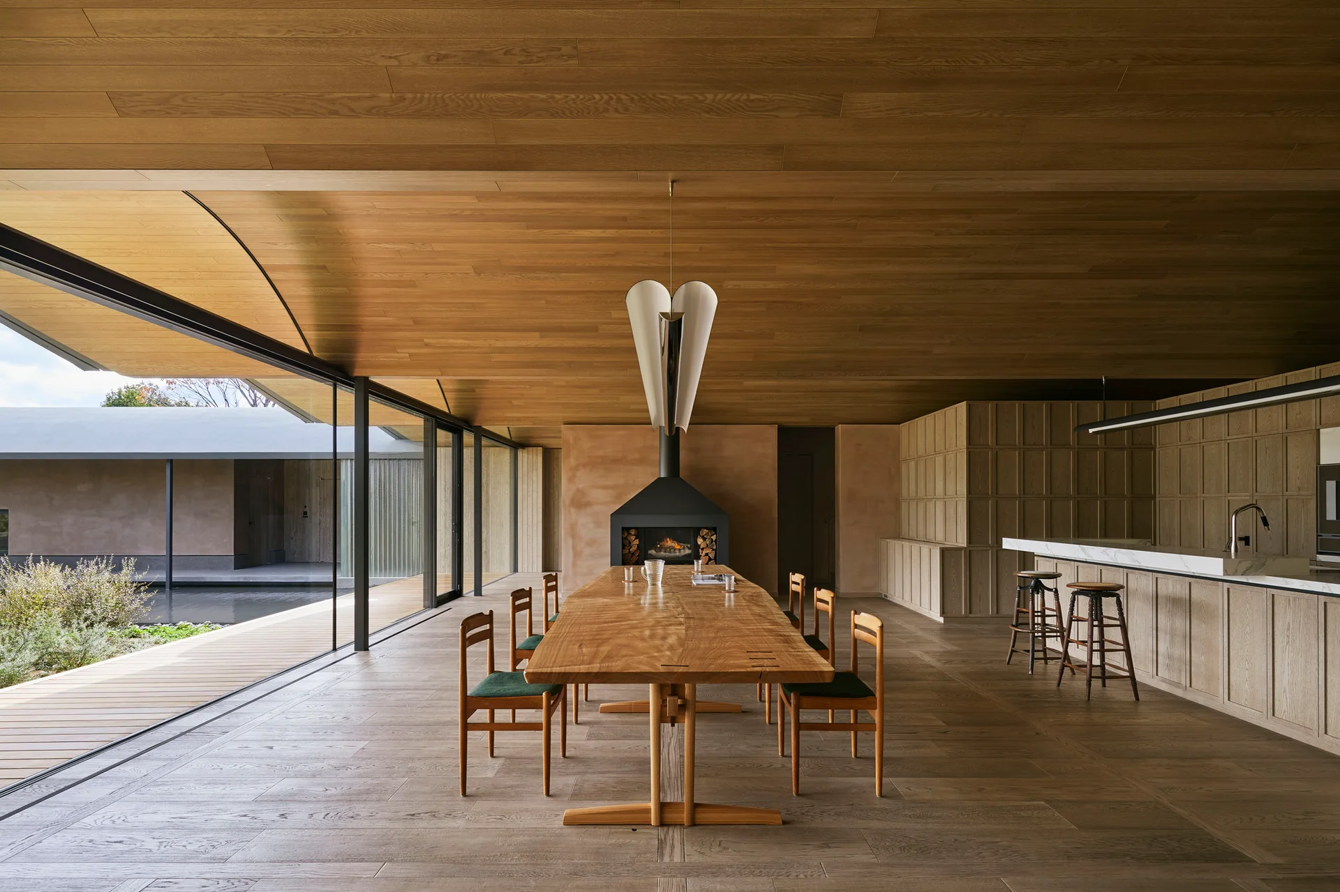 A minimalist dining area featuring a long wood table with green chairs, a wood plank ceiling, and sliding glass doors opening to a courtyard.