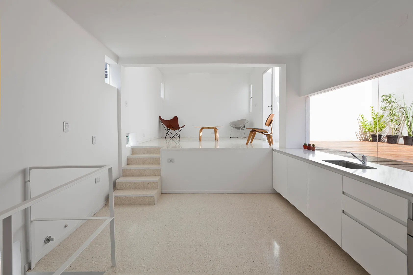 A minimalist split-level interior with a lower white kitchen and a raised living area containing a leather butterfly chair and a wooden coffee table.