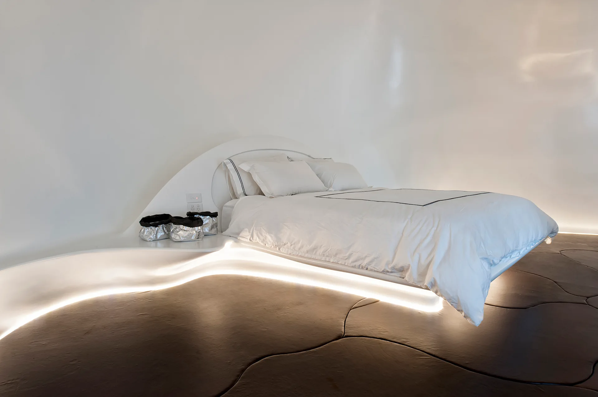 A minimalist white bedroom features a curved platform bed that floats above a dark segmented floor, highlighted by glowing under-bed lighting.