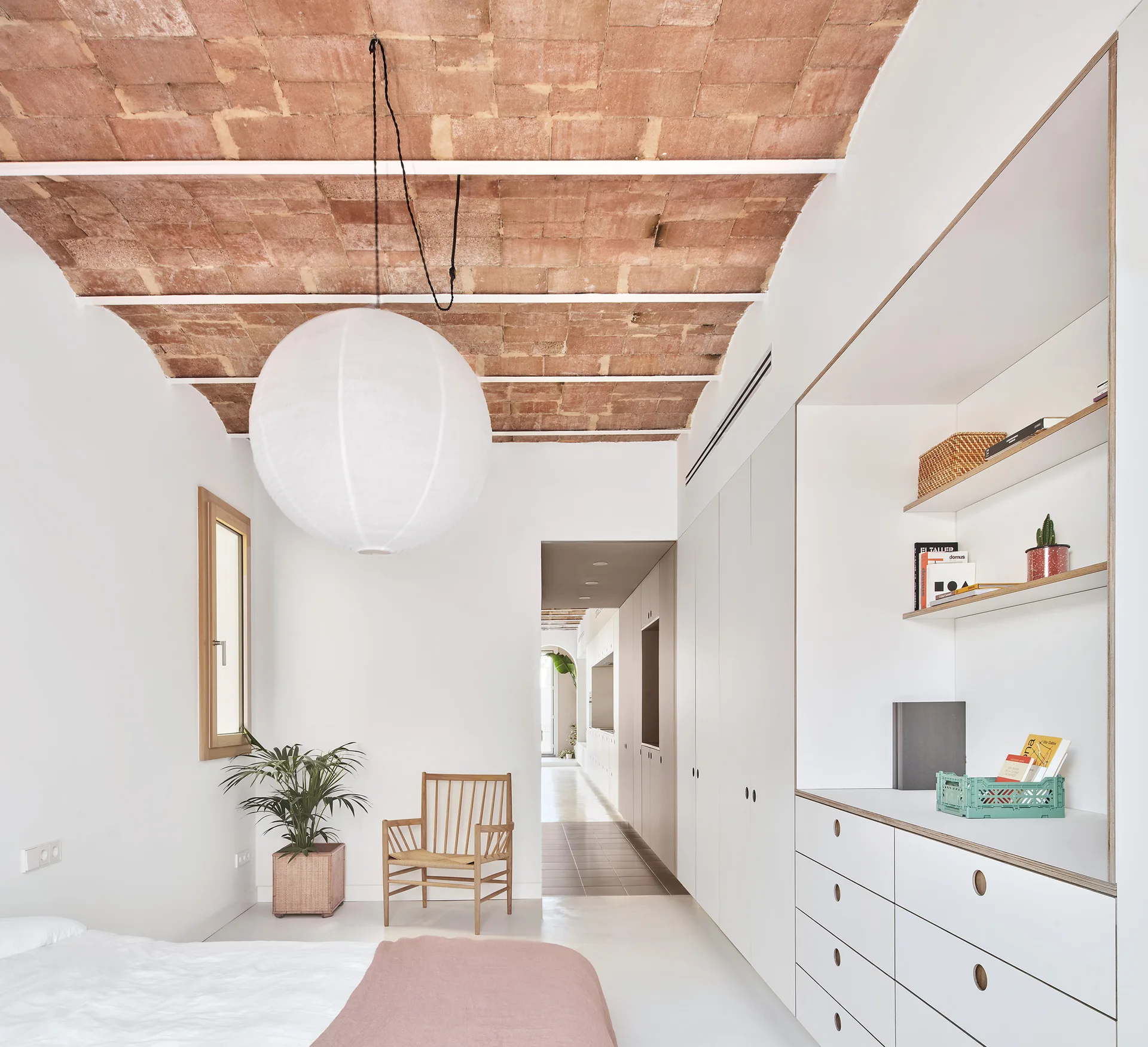 A minimalist bedroom featuring a terracotta vaulted brick ceiling, a large paper pendant light, and seamless white built-in storage cabinets.
