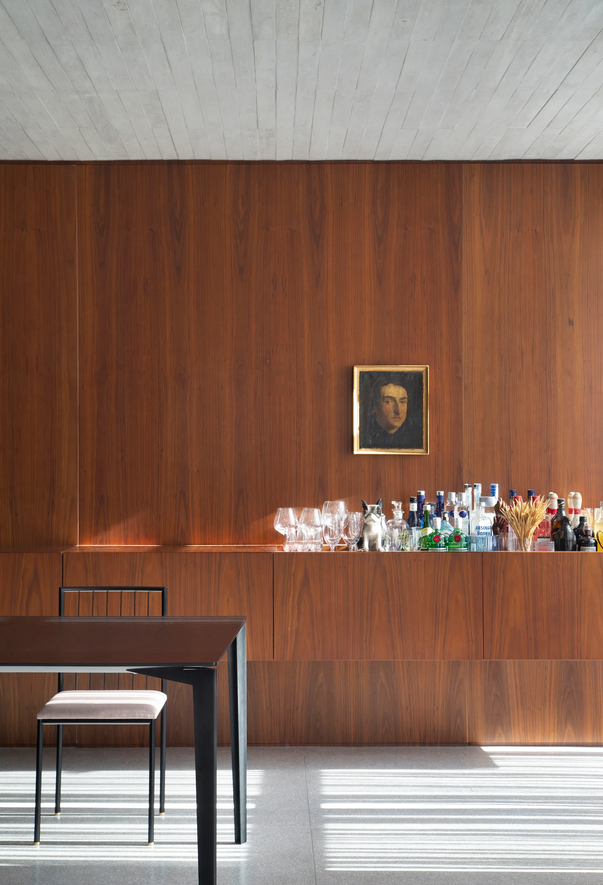 A modern dining room features a dark wood paneled wall with a built-in sideboard holding barware beneath a textured concrete ceiling.
