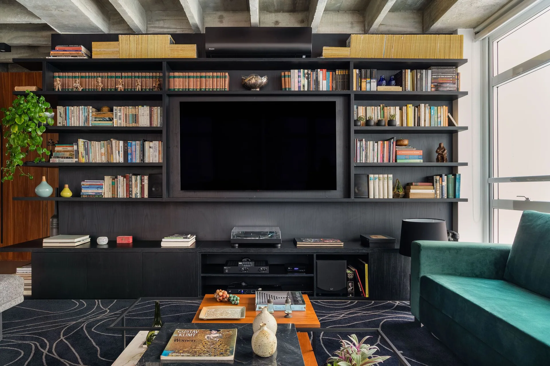 A modern living room featuring an expansive black built-in shelving unit, an exposed concrete ceiling, and a deep green sofa.