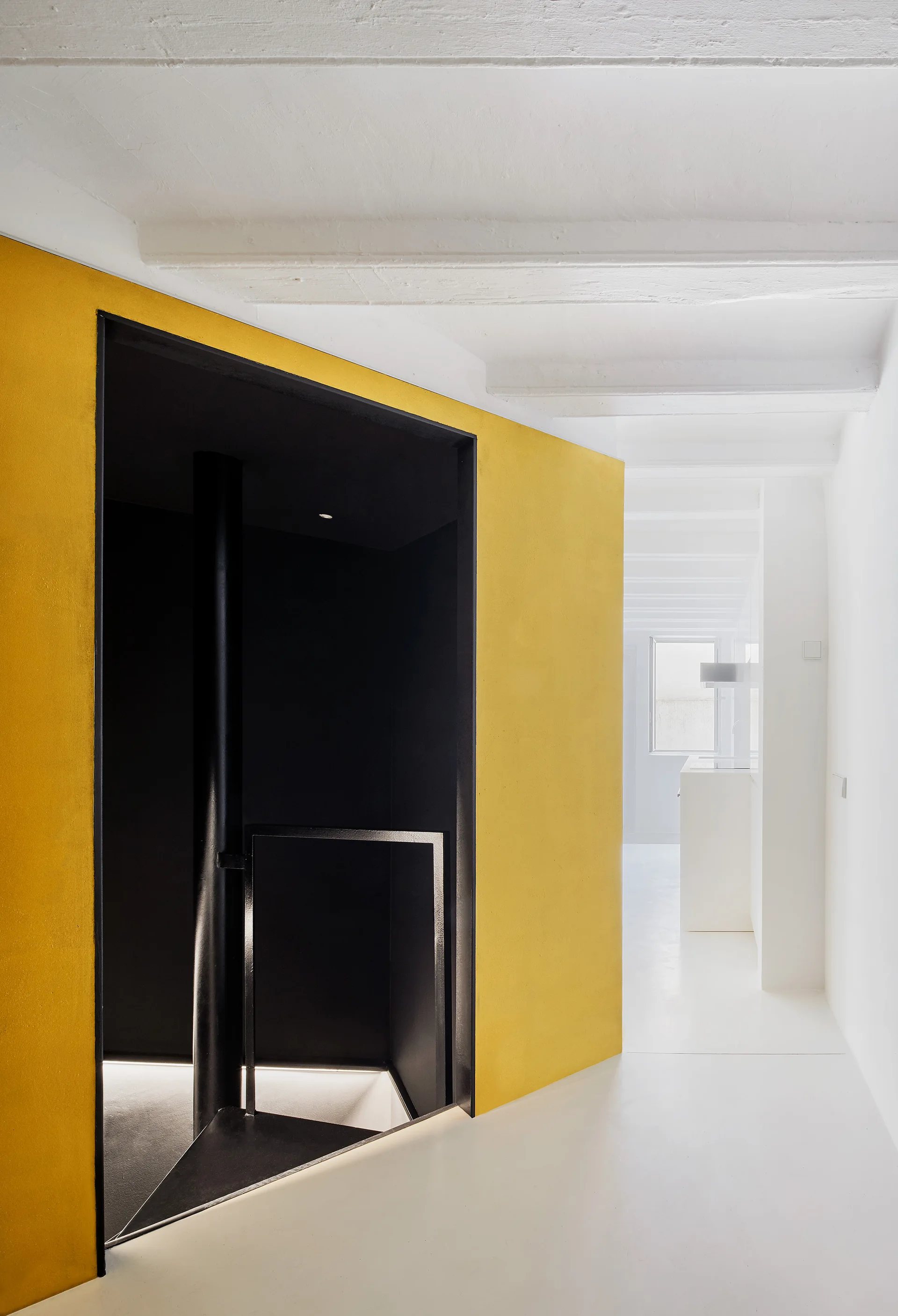 A minimalist white hallway featuring a bright yellow partition wall that frames a recessed black staircase entry beneath exposed ceiling beams.