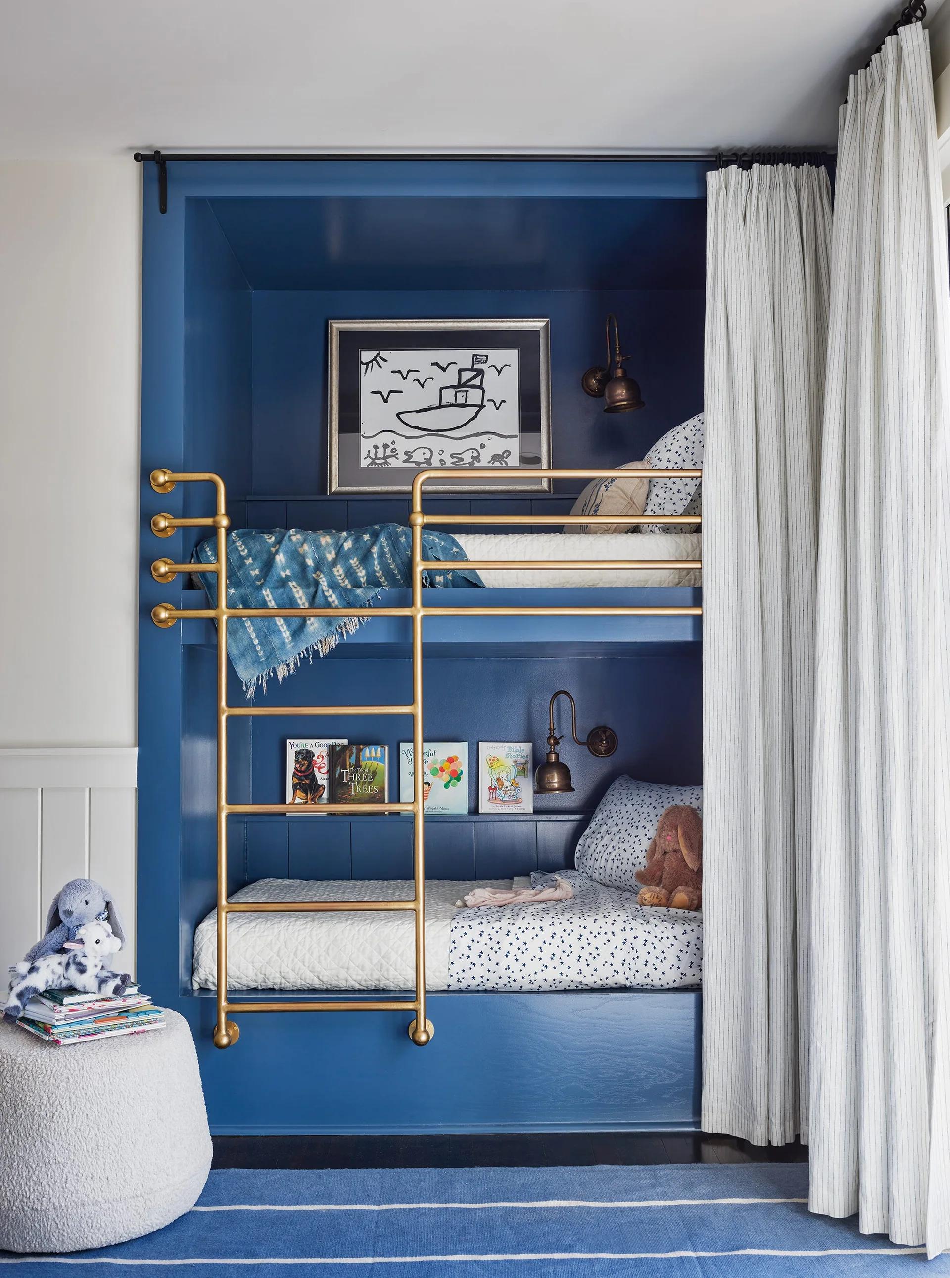 A built-in blue wooden bunk bed featuring a brass ladder, warm brass sconces, integrated book shelves, and striped curtains in a kids bedroom.