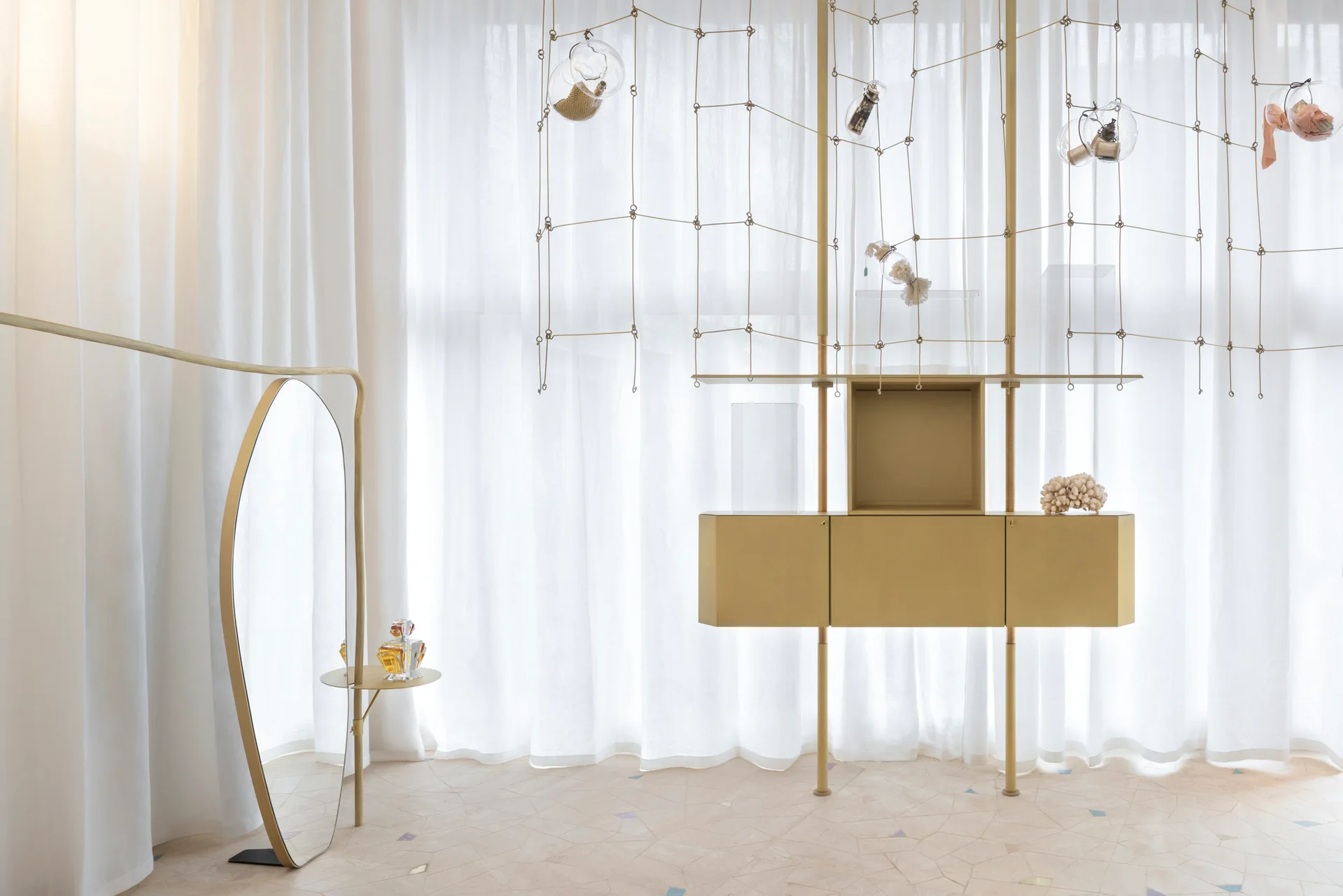 A gold metal shelving unit and a matching floor mirror stand on a terrazzo floor in front of floor-to-ceiling sheer white curtains.