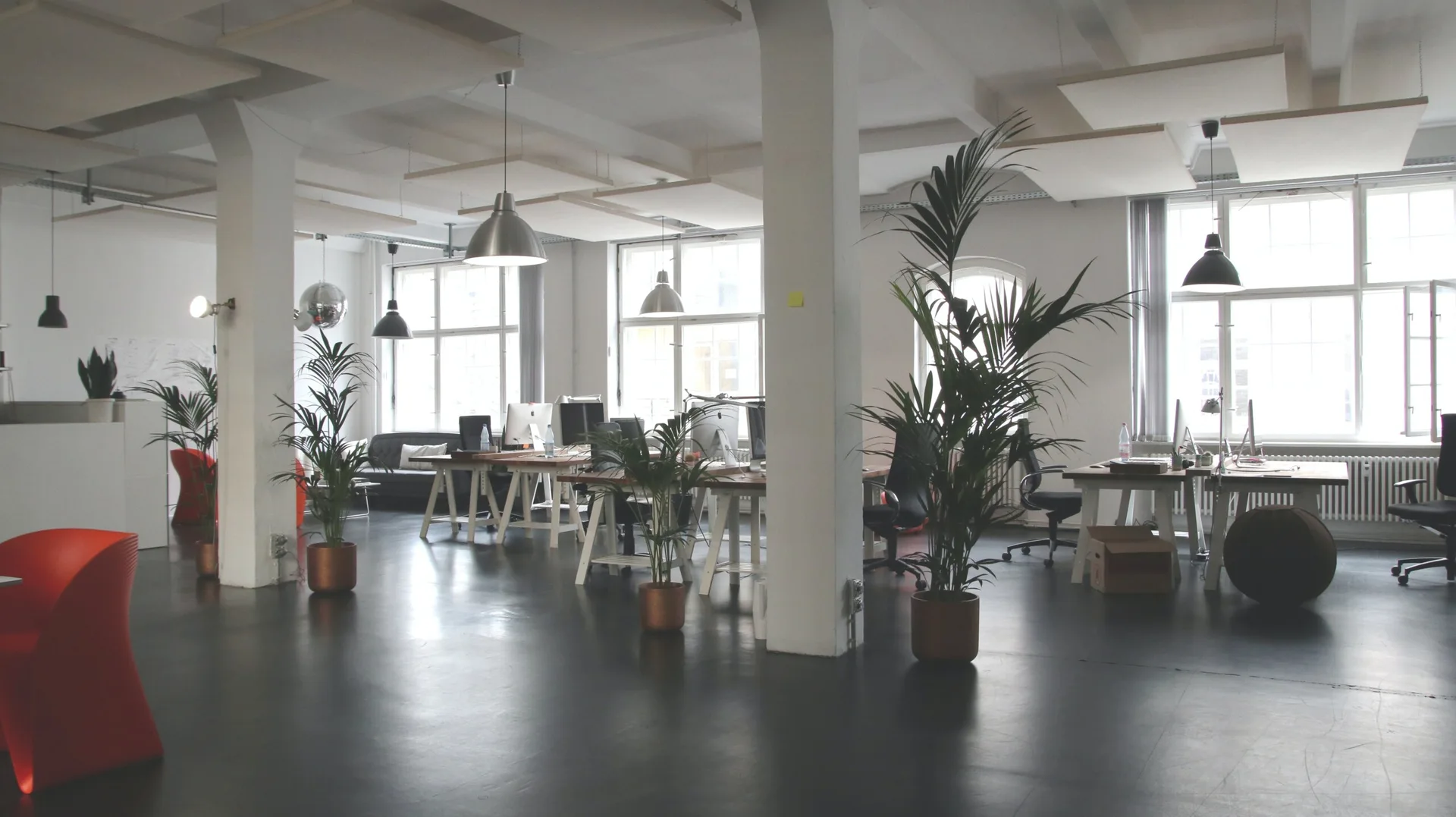 An open-plan office space featuring white structural columns, dark floors, wooden desks, metal pendant lights, and tall potted palms.