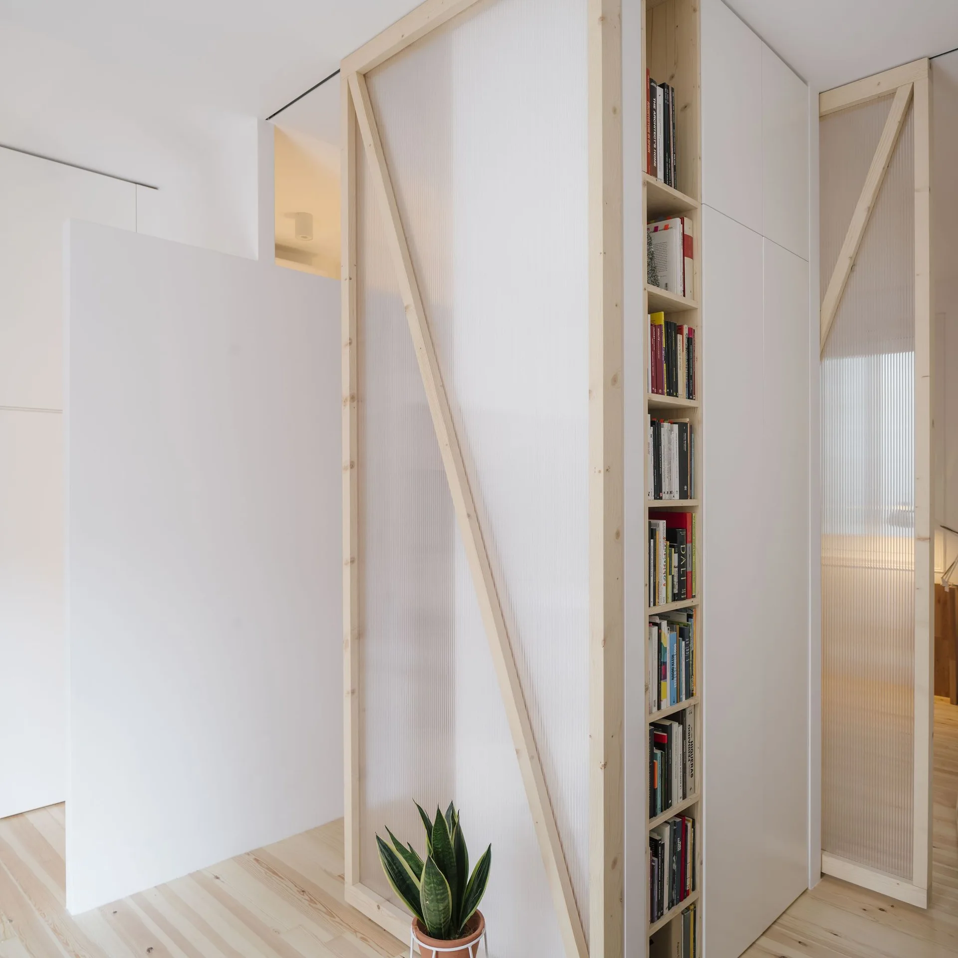 A light wood-framed partition wall with translucent fluted panels stands on a light-colored wood floor next to a white built-in bookcase.