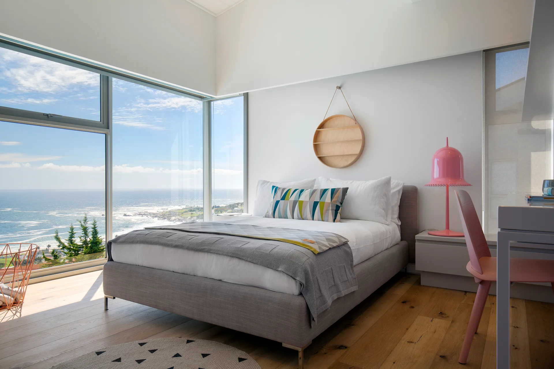 A modern bedroom features a gray upholstered bed next to a large corner window showing an ocean view, with a pink chair and lamp nearby.