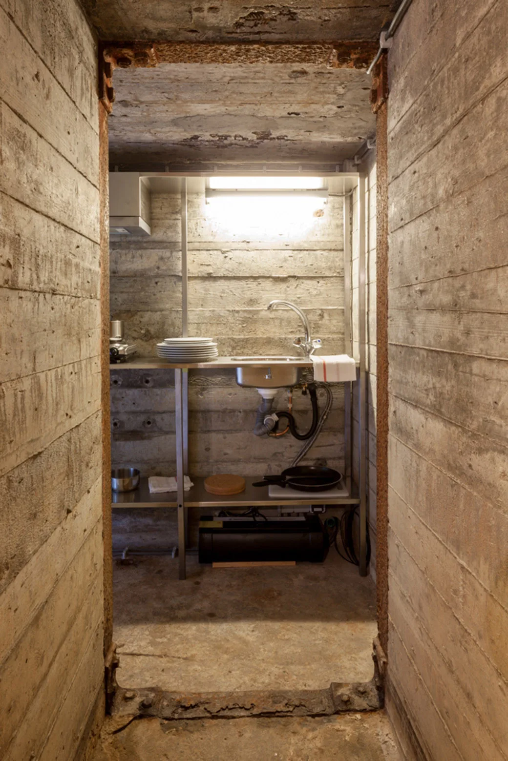 A compact industrial kitchen with a stainless steel sink unit is framed by a rusted metal doorway and raw concrete walls.