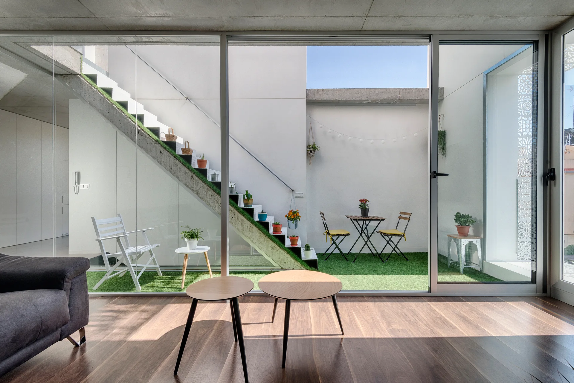 A living room with dark wood flooring looks through a sliding glass door to an outdoor patio with artificial turf and a plant-decorated concrete staircase.