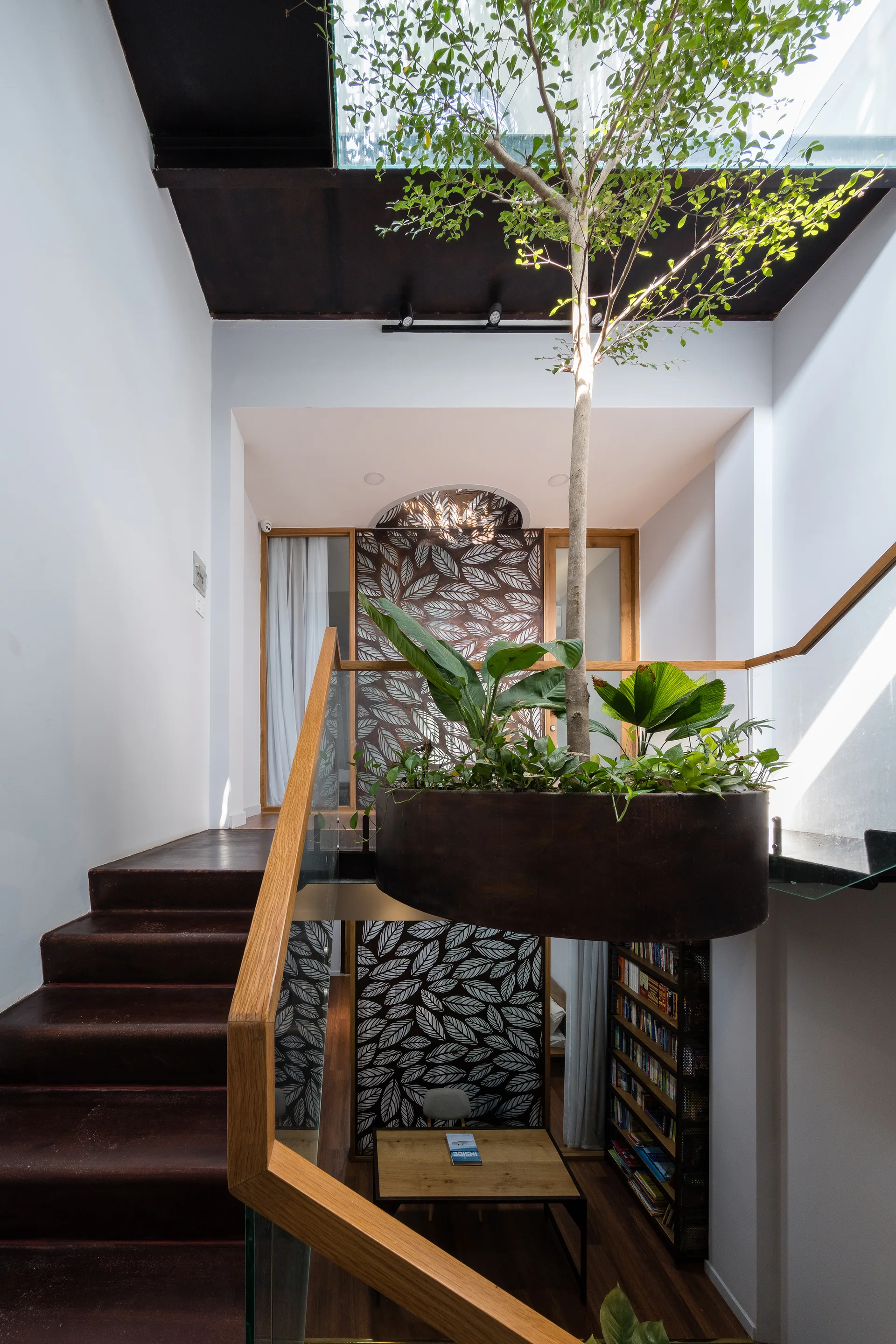 A modern staircase with dark treads and a glass railing wraps around a suspended metal planter holding a tall indoor tree beneath a skylight.