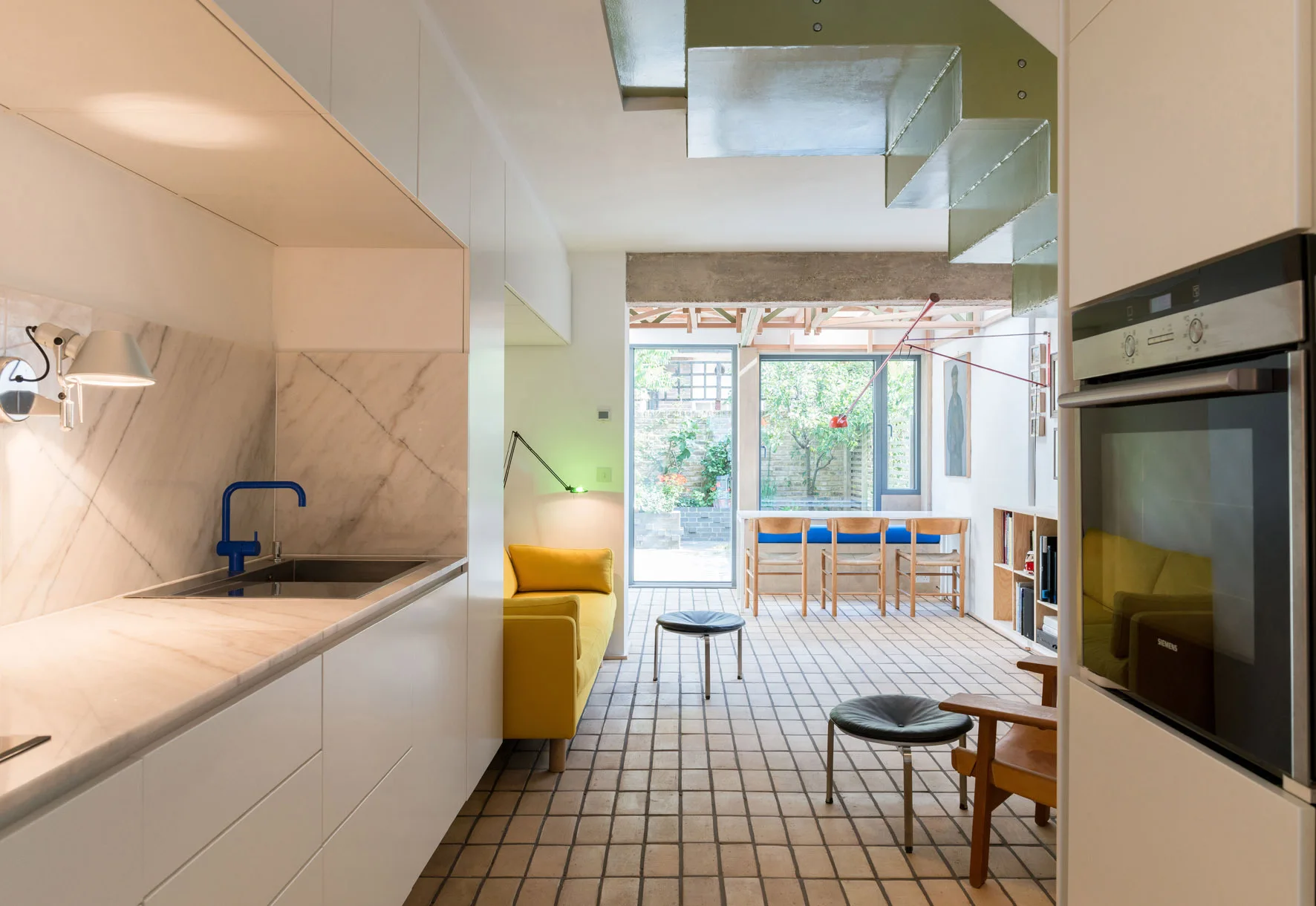 An open-plan residential interior showing a white minimalist kitchen with a blue faucet, a yellow sofa, and grid-patterned floor tiles.