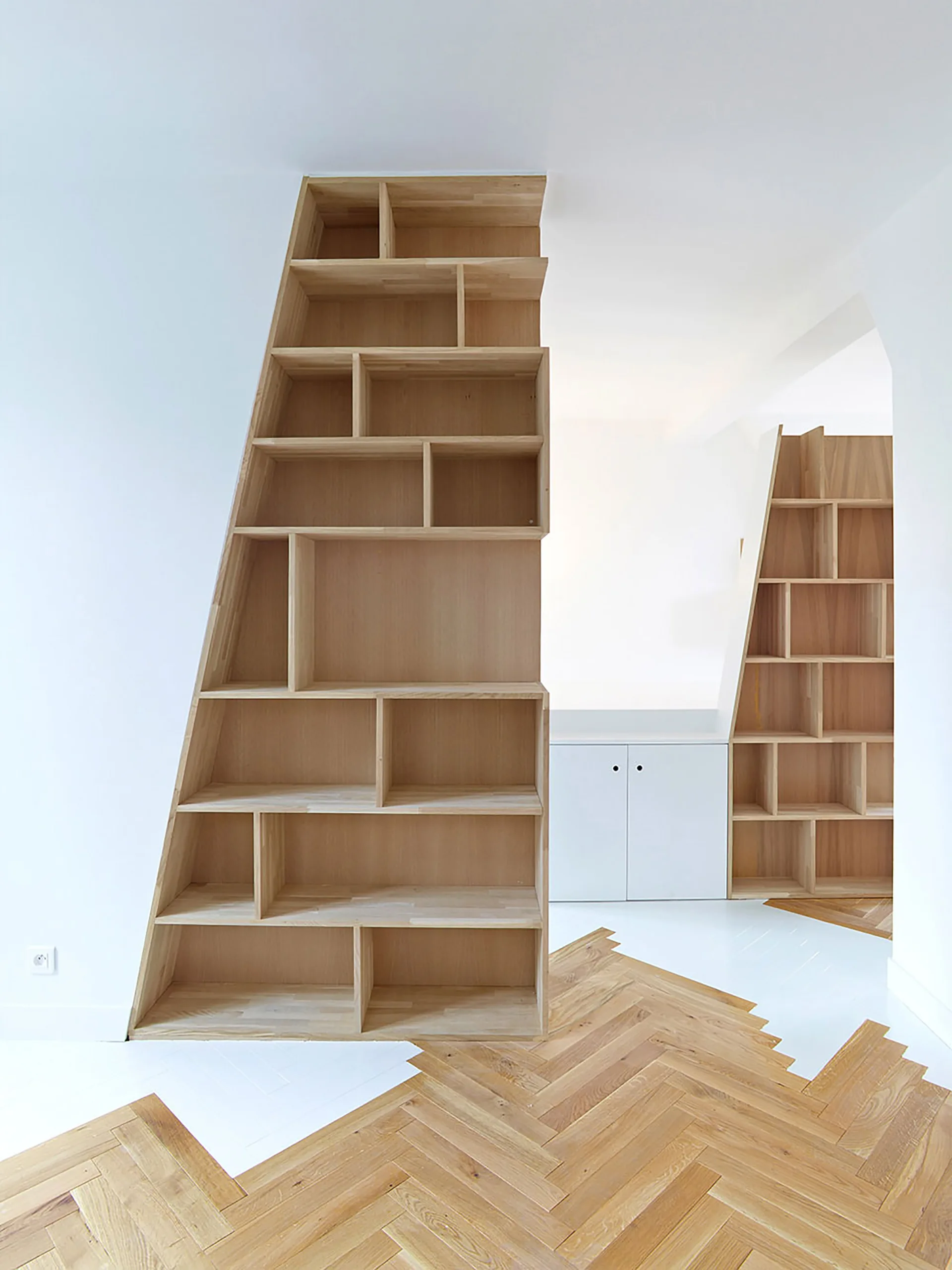 Two custom slanted wooden bookshelves stand on a floor featuring a graphic transition between oak herringbone parquet and white painted planks.