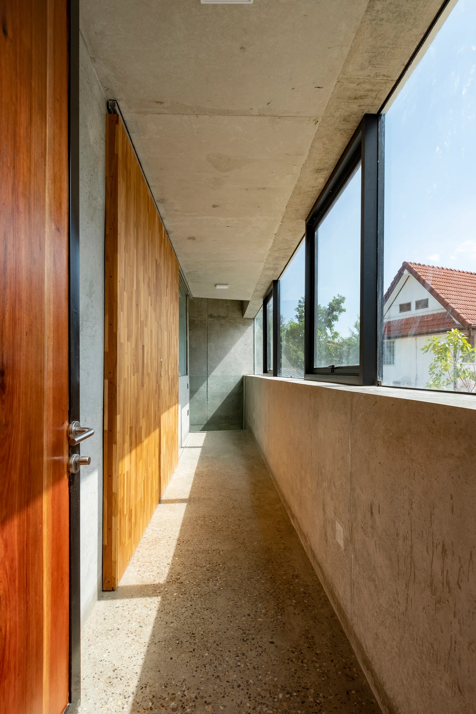 A narrow concrete hallway featuring a warm wood sliding door, a speckled terrazzo floor, and a long wall of large windows.