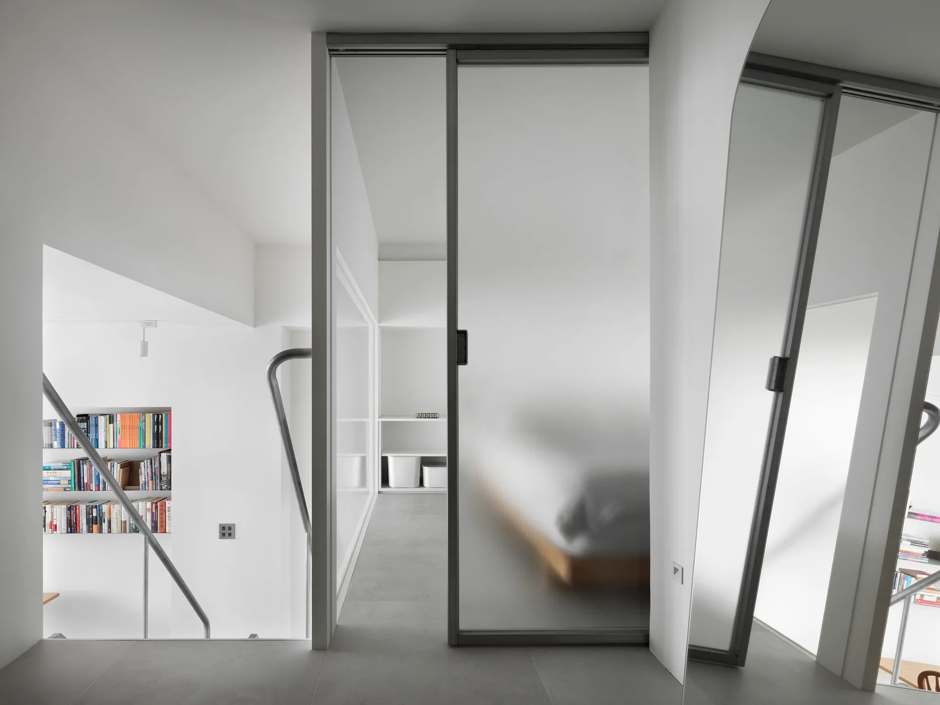 A minimalist stairwell landing features a frosted glass sliding door next to a metal handrail and a recessed bookshelf.