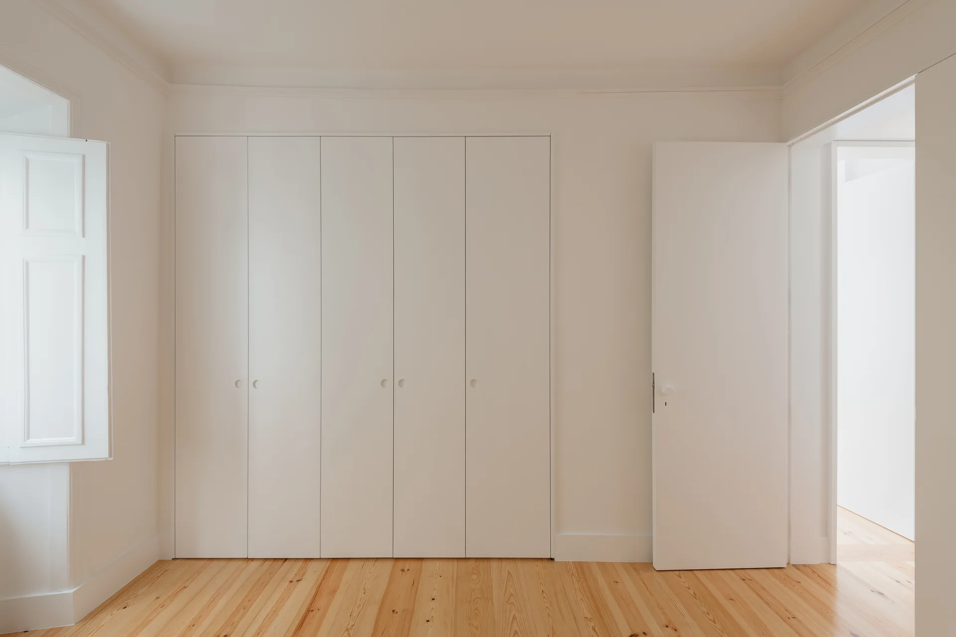 A minimalist off-white built-in closet with clean vertical lines stands next to an open door on light wood flooring.