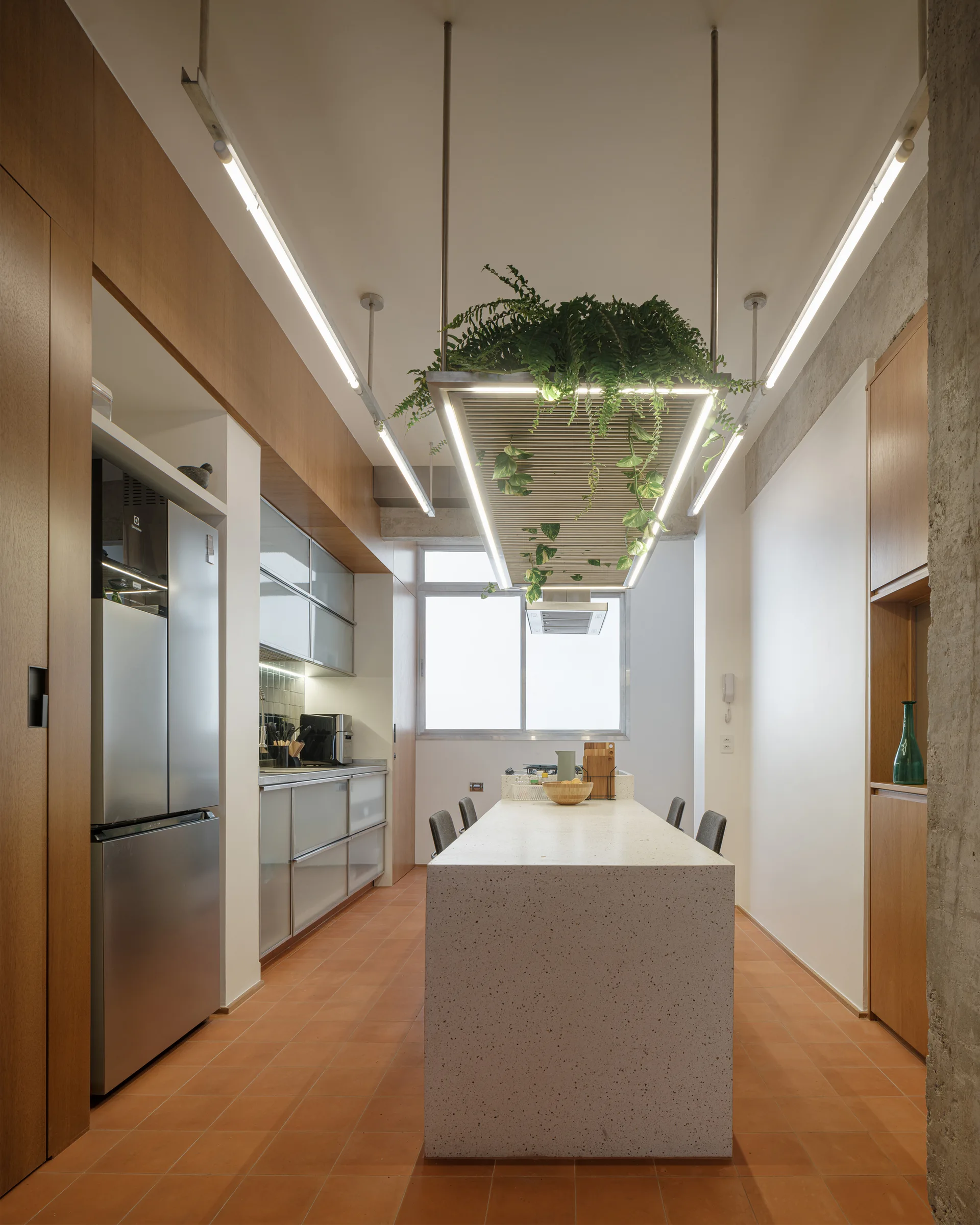 A modern galley kitchen featuring a central terrazzo island, terracotta tile flooring, wood cabinetry, and a suspended planter light fixture.
