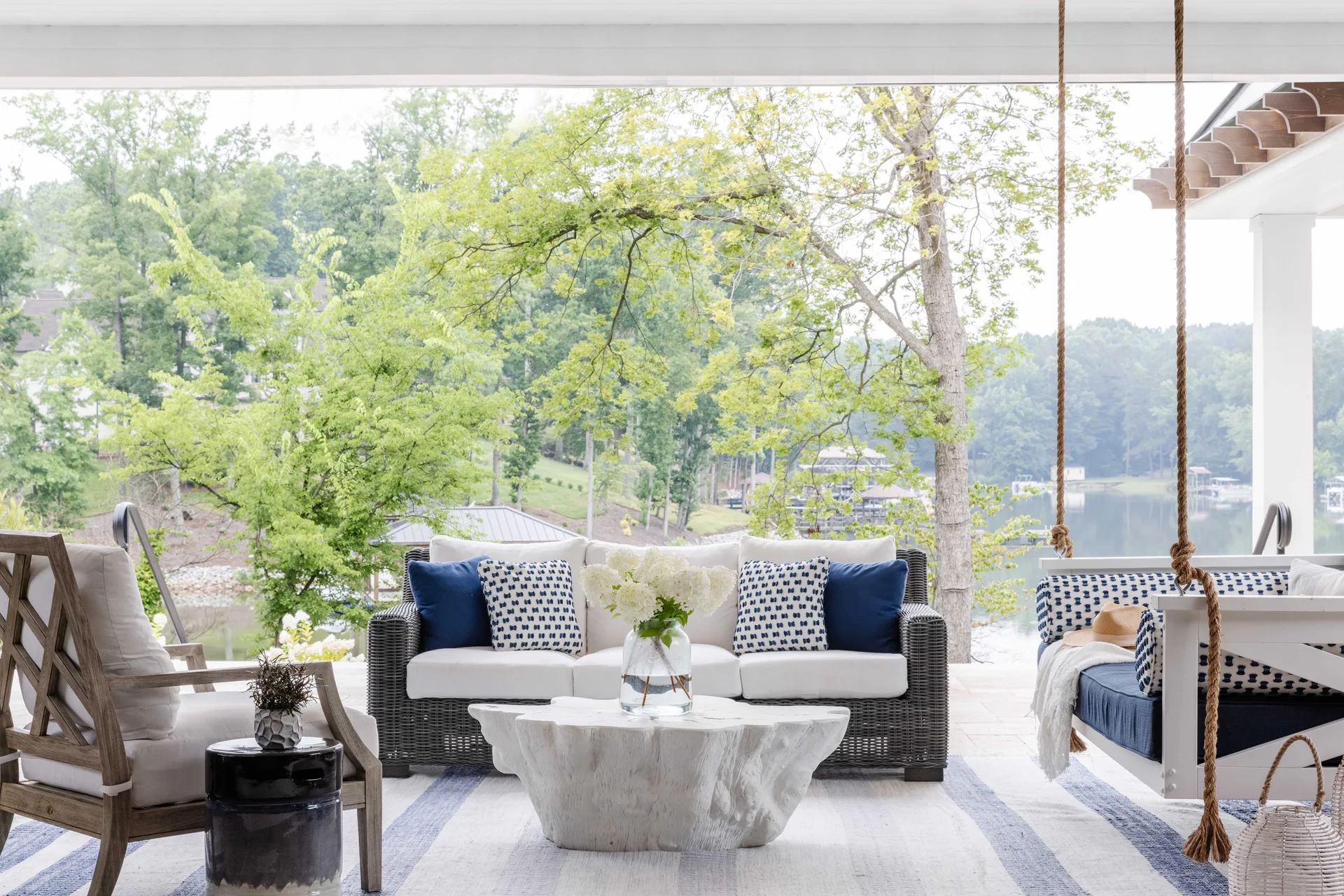A covered outdoor porch features a wicker sofa, a hanging swing bench, and an organic-shaped white coffee table overlooking a lake.
