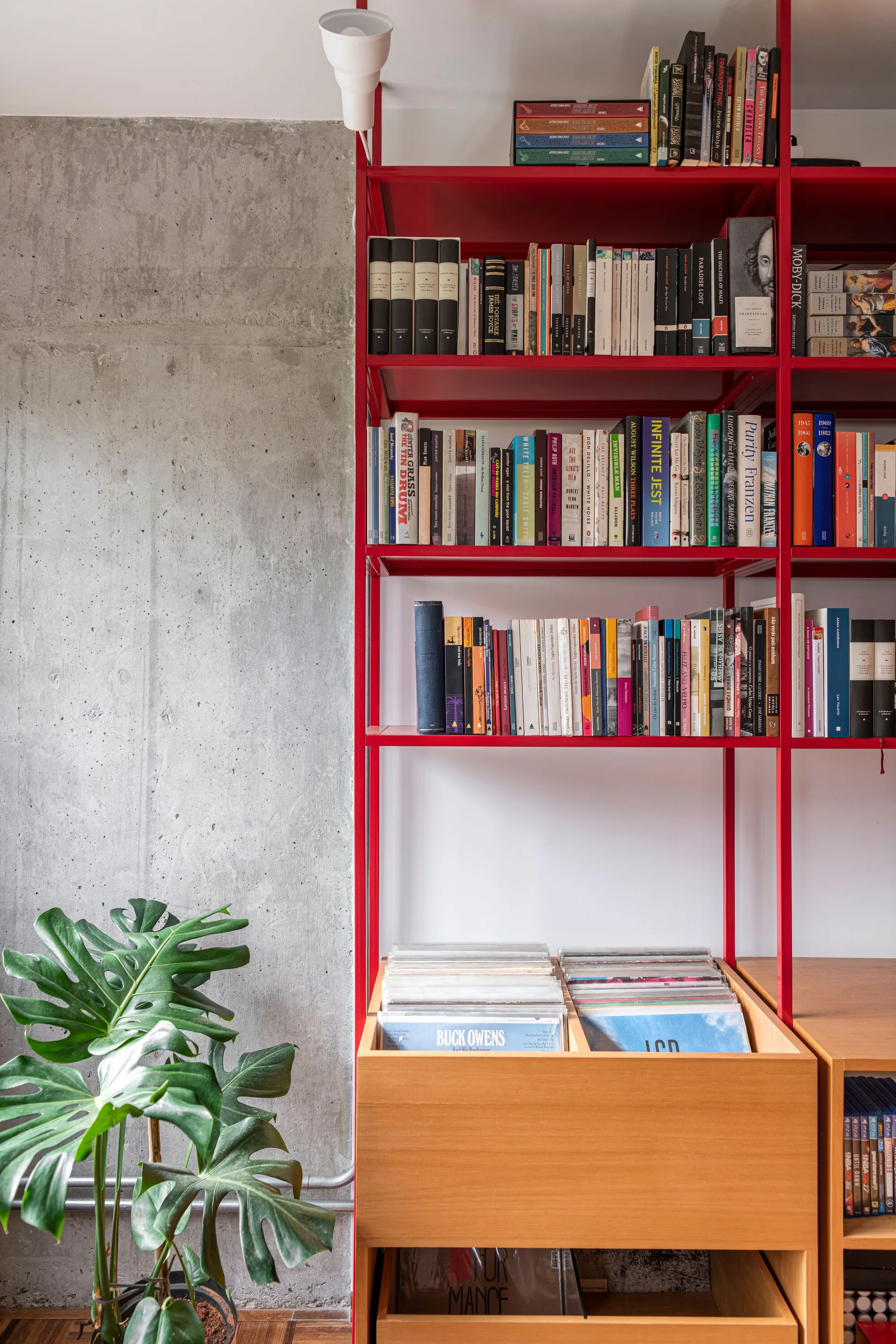 A red metal open shelving unit filled with books and vinyl records stands next to a raw concrete wall and a green monstera plant.
