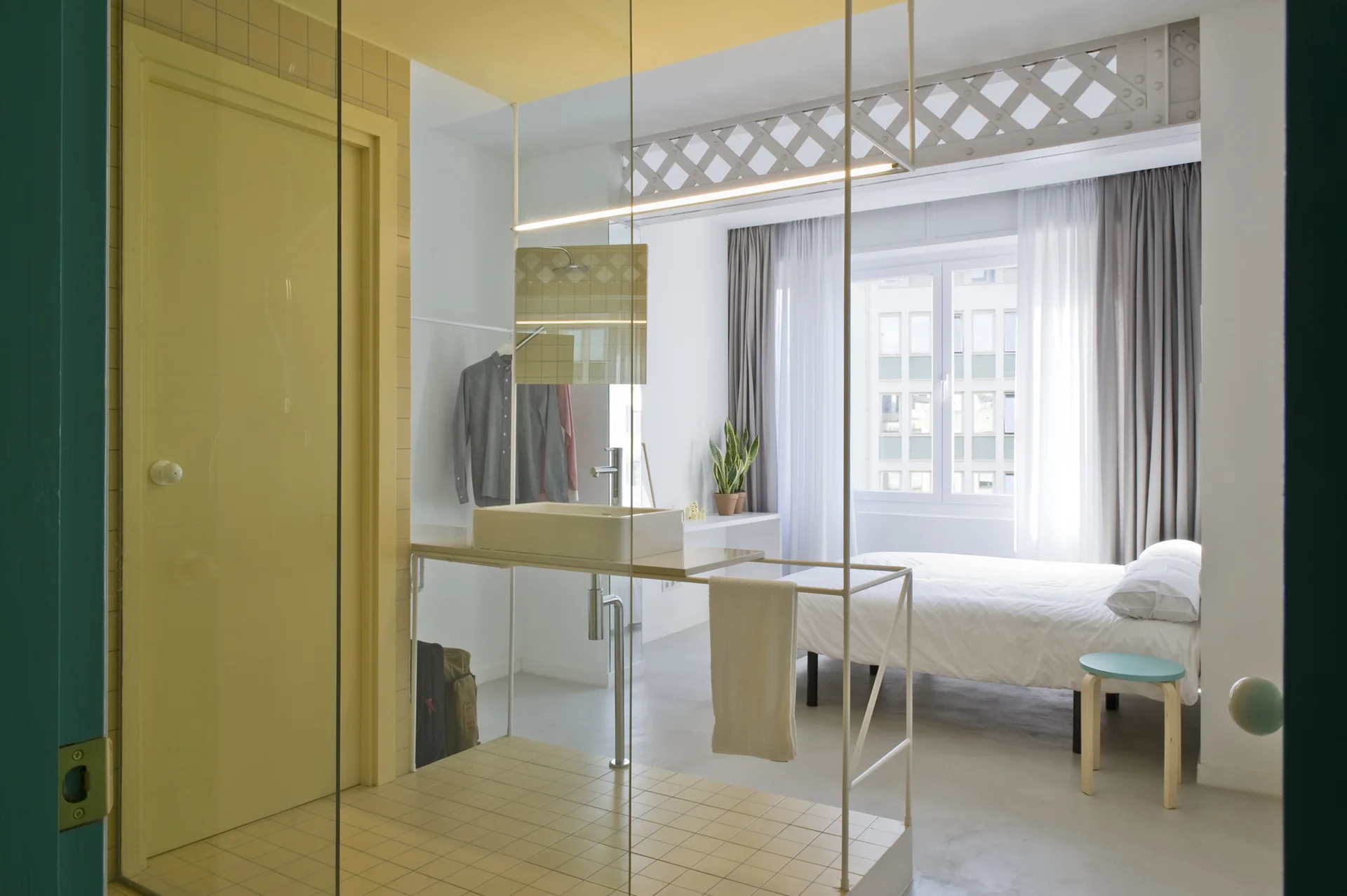 A minimalist bedroom features a glass-partitioned bathroom vanity area with yellow tiling, a white bed, and light grey floors.
