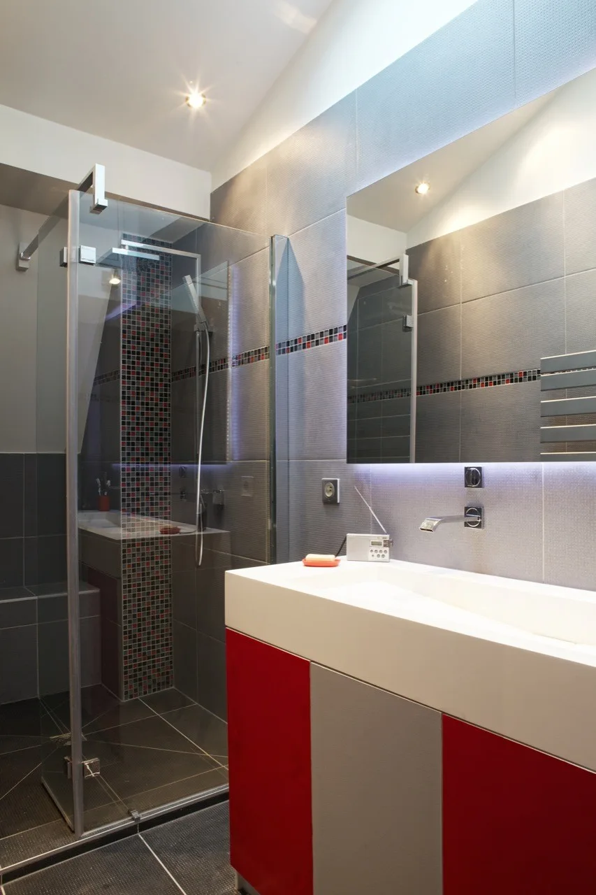 A modern bathroom featuring a red and grey color-blocked vanity, a glass shower enclosure with red mosaic tiles, and large grey wall tiles.