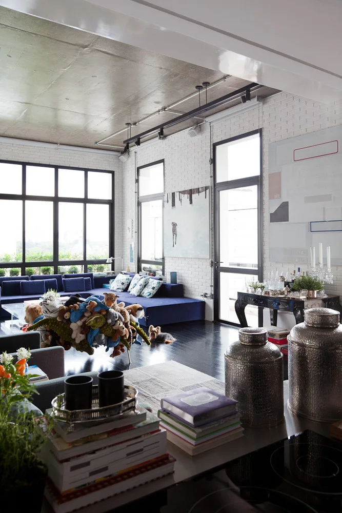 An industrial loft living room with white painted brick walls, a large blue sectional sofa, black window frames, and glossy dark wood floors.