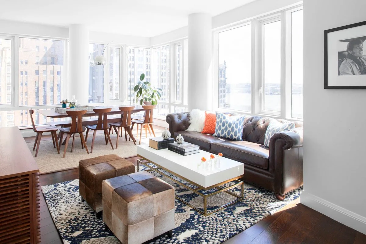 A bright open-concept living room features a brown tufted leather sofa, a white coffee table, cowhide ottomans, and large windows with city views.