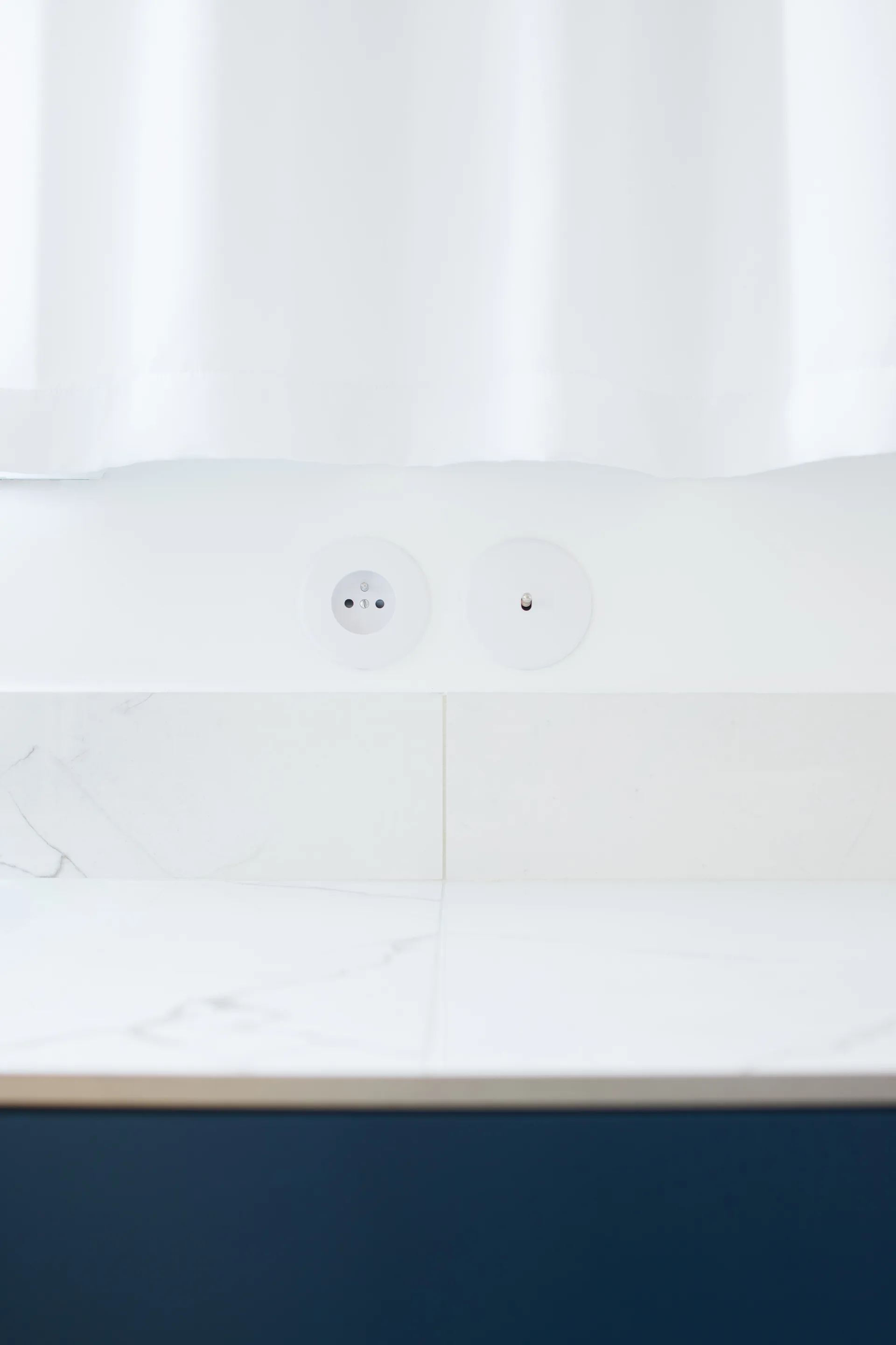 Minimalist circular white electrical outlets set into a white wall above a veined marble backsplash and a dark blue cabinet base.