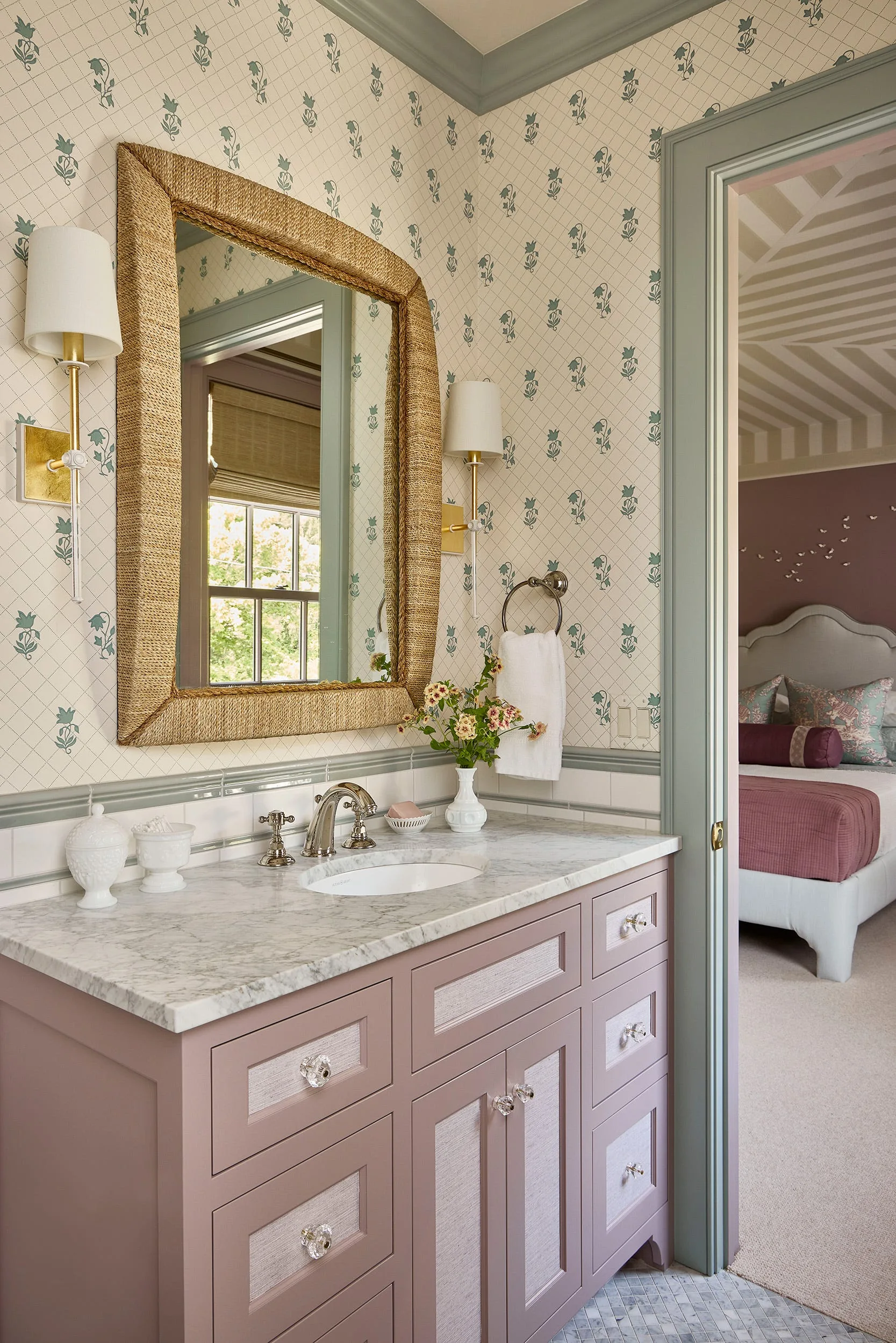 A bathroom features a mauve vanity with a marble countertop, floral trellis wallpaper, a woven fiber mirror, and a view into an adjacent bedroom.