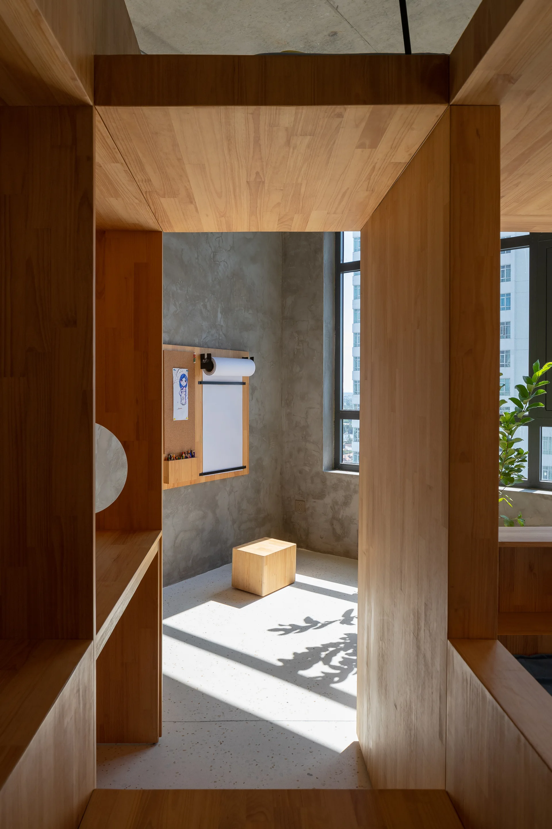 A minimalist kids' play nook viewed through a custom wooden framing structure, featuring concrete walls, a small wood stool, and a terrazzo floor.