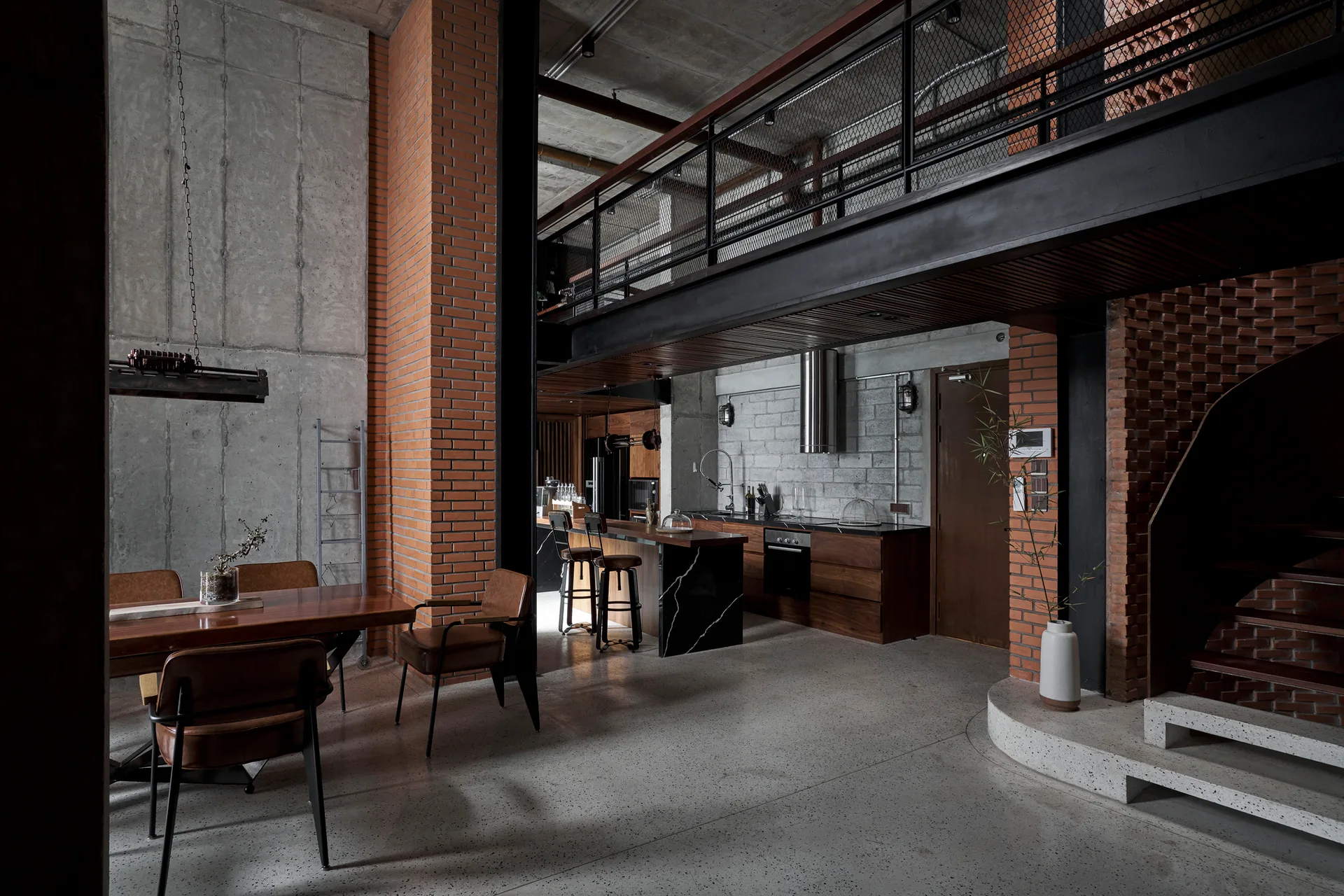 An industrial loft kitchen and dining space with a wooden table, polished concrete floor, exposed brick pillars, and a metal mezzanine balcony.