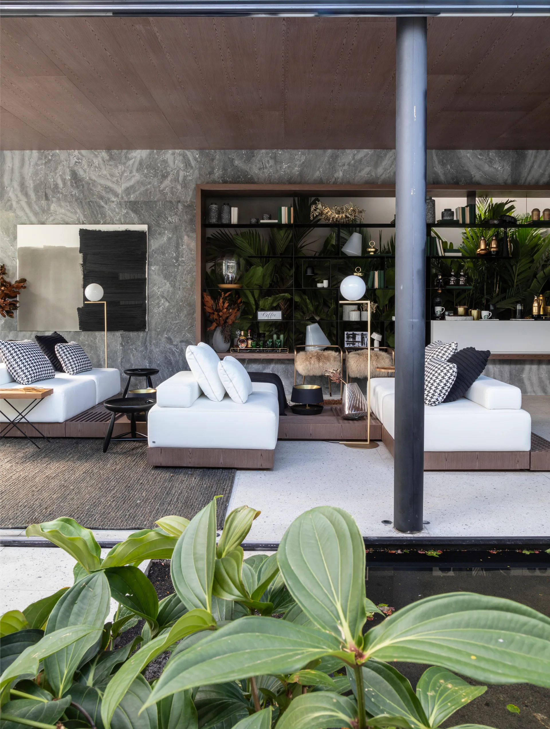 A covered patio features white modular sofas, a grey stone wall, and a recessed shelving unit filled with potted leafy green plants.