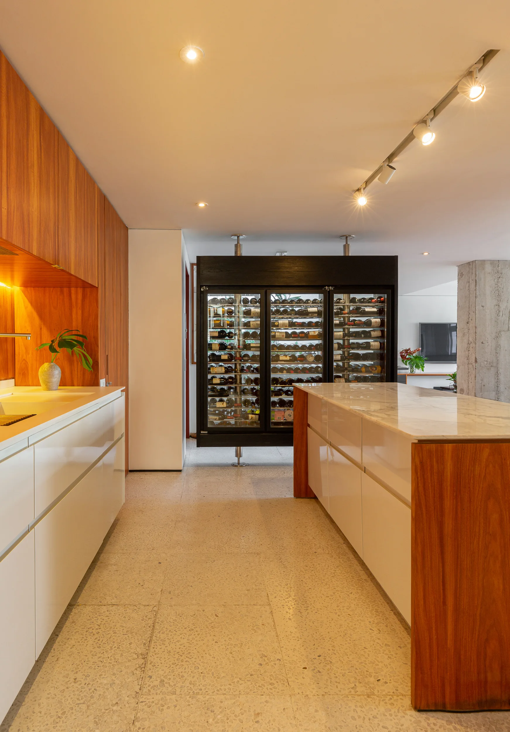 A modern kitchen featuring a marble-topped island, white cabinetry with wood accents, speckled terrazzo flooring, and a large integrated wine refrigerator.