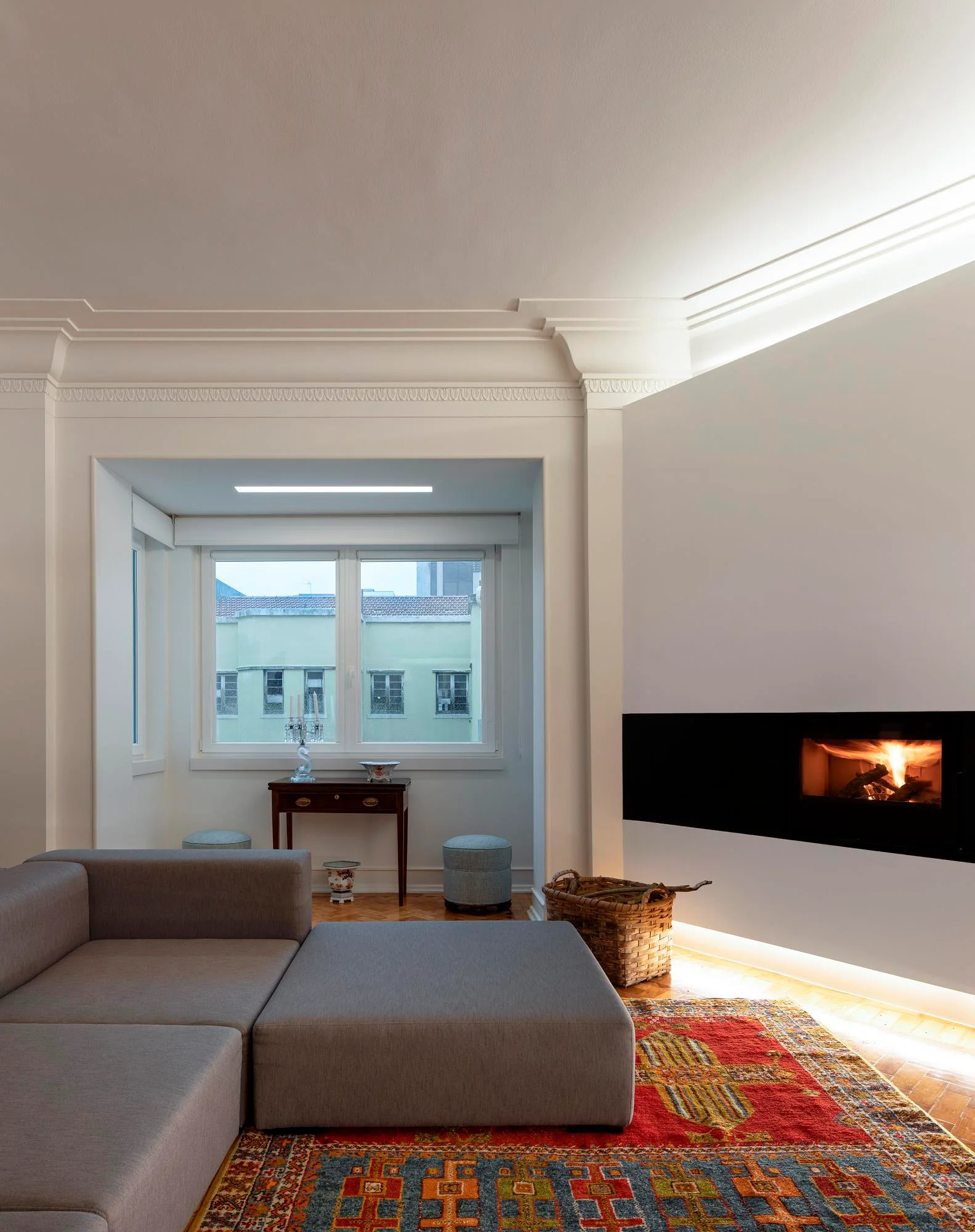 A modern grey sectional sofa sits on a colorful patterned rug next to a floating fireplace wall under traditional white crown molding.