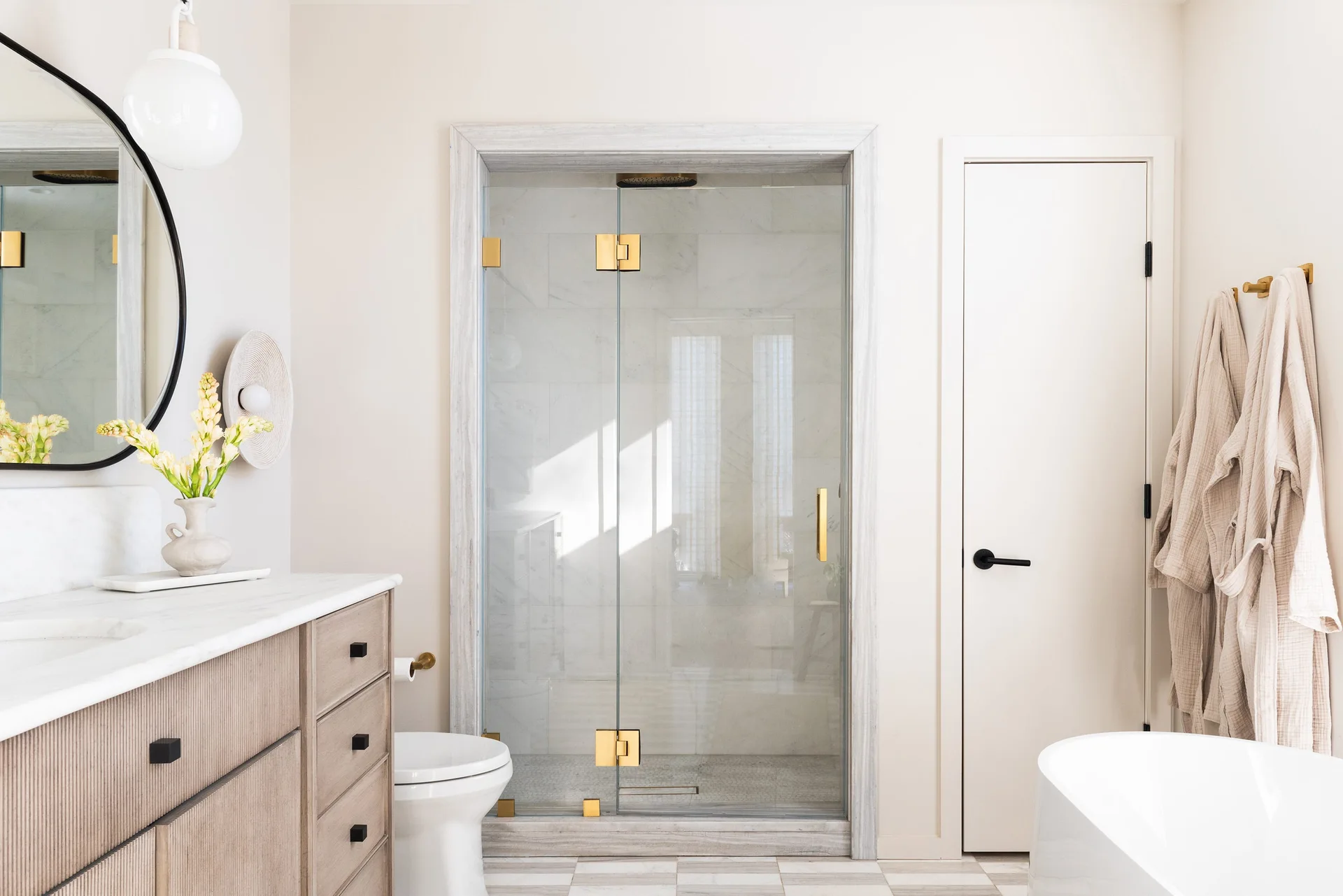 A bright bathroom features a fluted wood vanity with a marble top, a glass shower with brass hardware, and a tiled checkerboard floor.