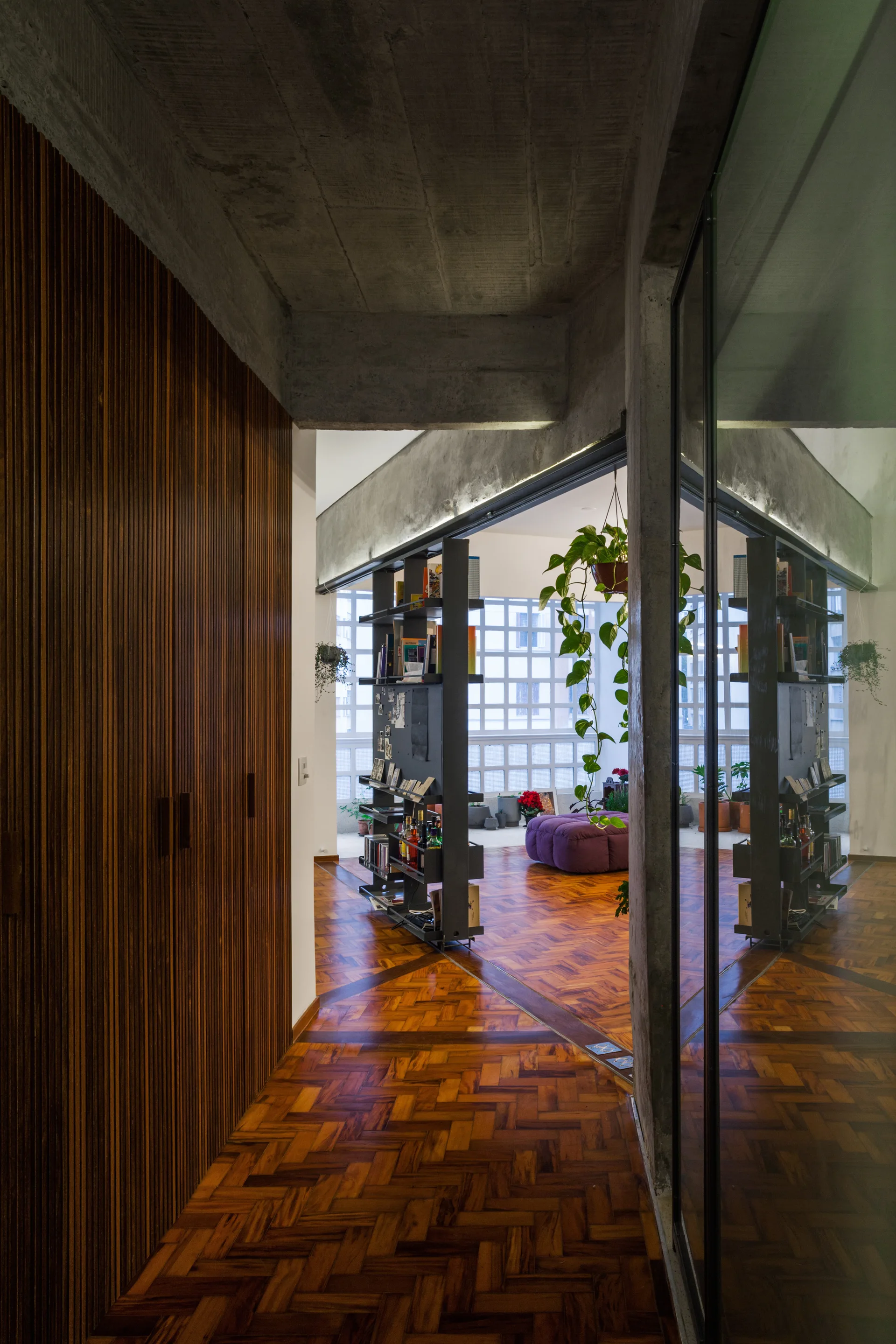 A hallway featuring vertical wood-slat wall paneling, herringbone parquet flooring, and an exposed raw concrete ceiling leading into a living room.