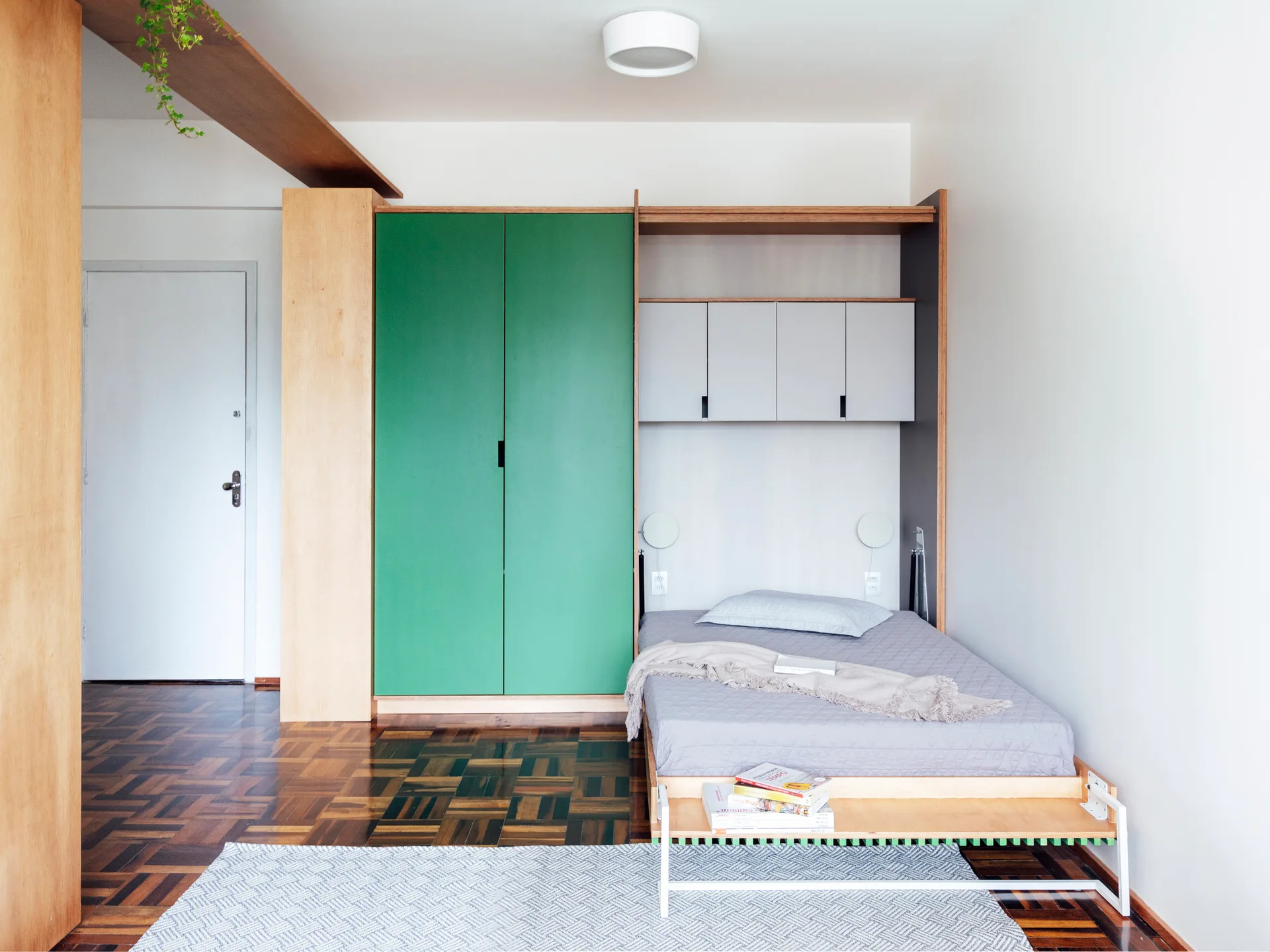 A small bedroom features parquet wood floors, a built-in wooden storage unit with green closet doors, and a low-profile bed tucked into a niche.