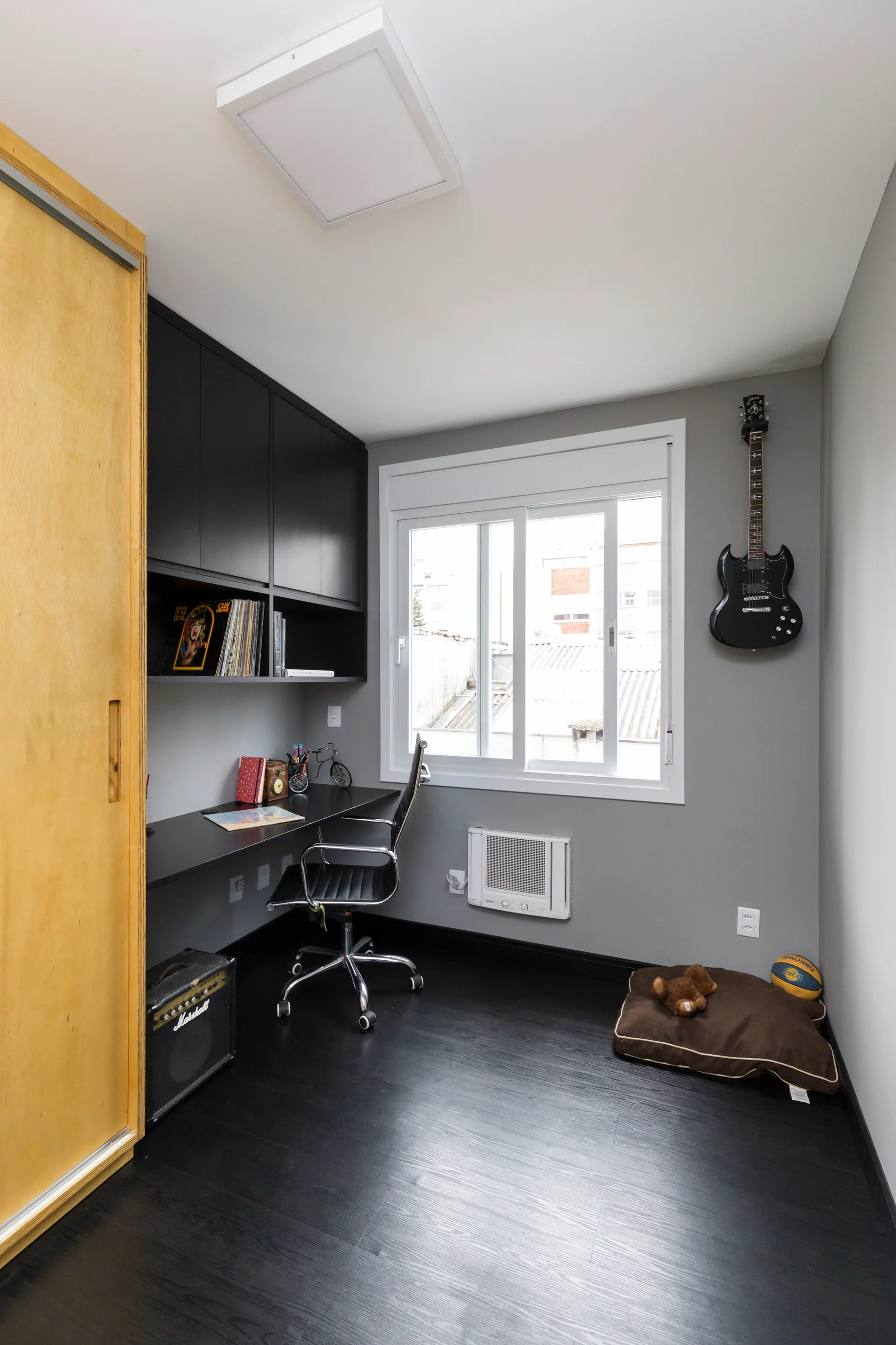 A small home office featuring gray walls, a black built-in desk with overhead cabinets, an office chair, and a light wood sliding closet door.