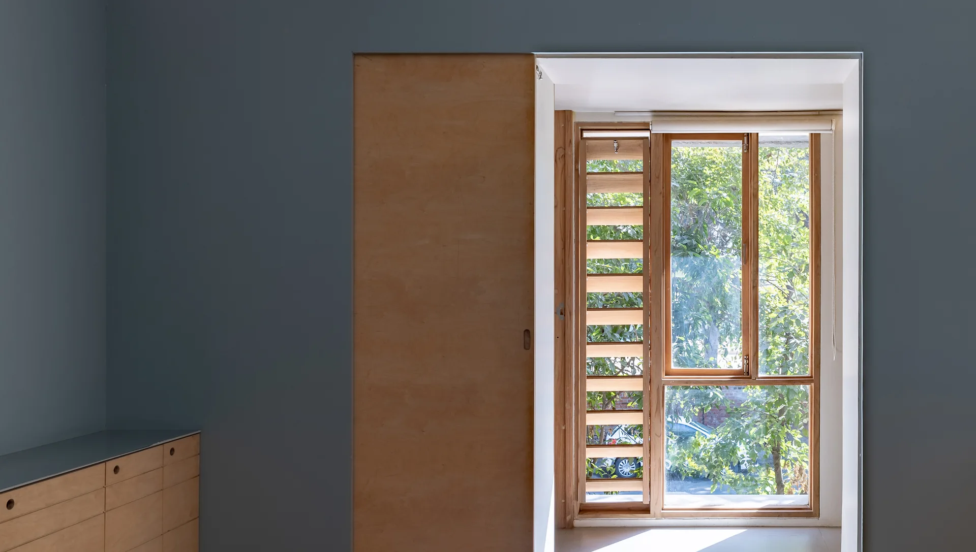 A light plywood sliding door sits open next to a wood-framed window with integrated louvers against a solid blue-grey wall.