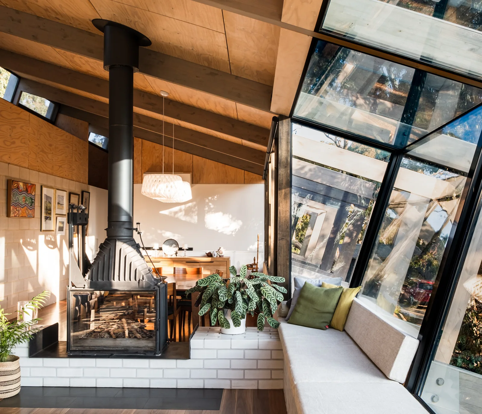 A modern living area with a sloped wooden ceiling, a freestanding black fireplace on a white brick hearth, and a built-in bench next to glass windows.