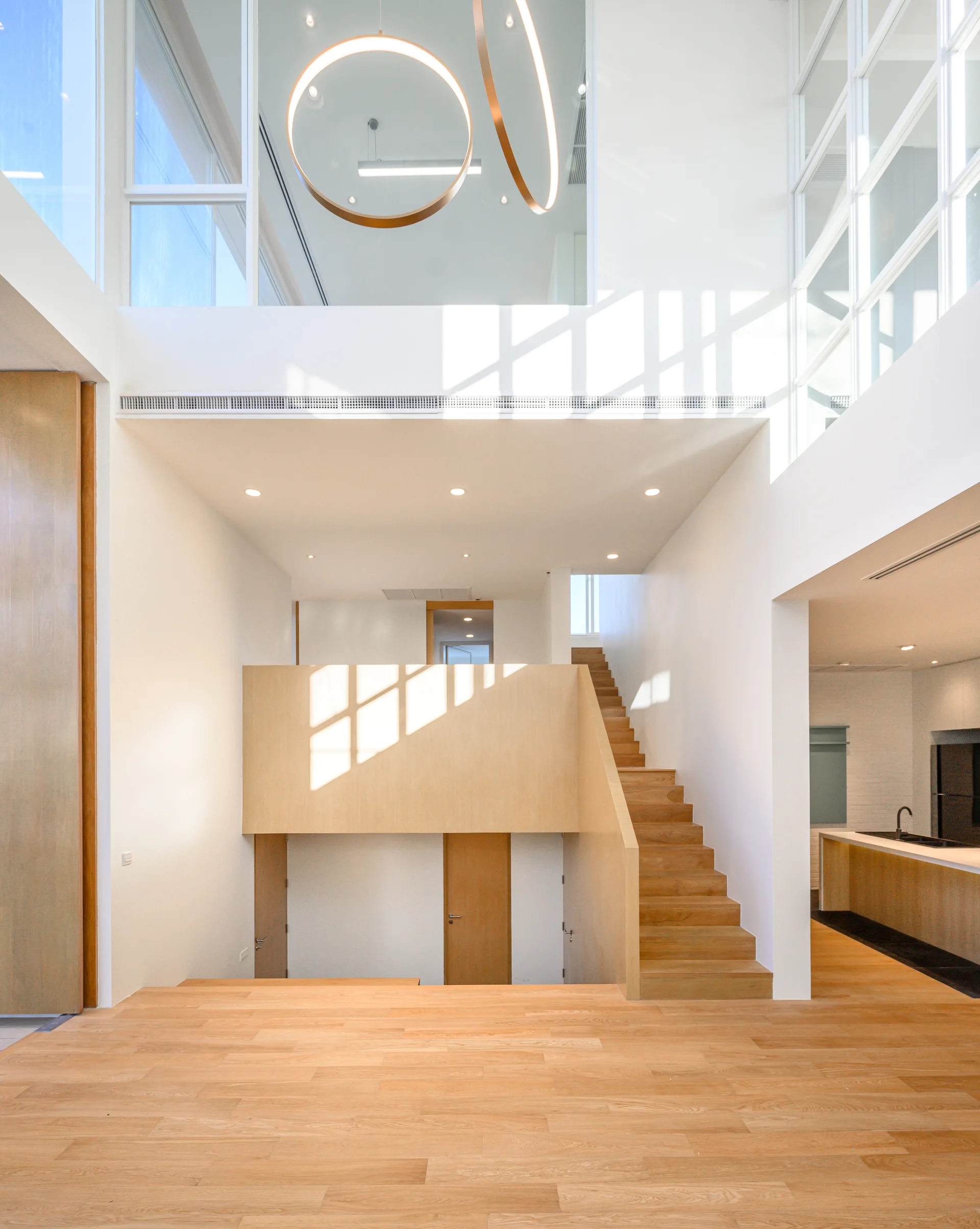 A bright minimalist home interior features a light wood staircase, matching floors, white walls, and circular brass ring pendant lights.