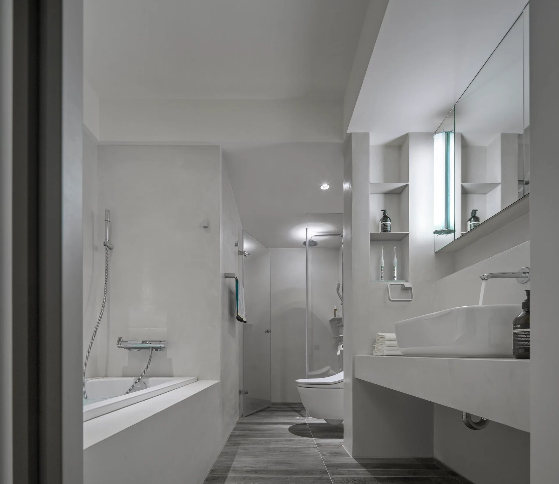 A minimalist white bathroom features a built-in tub, a white floating vanity with a vessel sink, and a recessed wall niche.