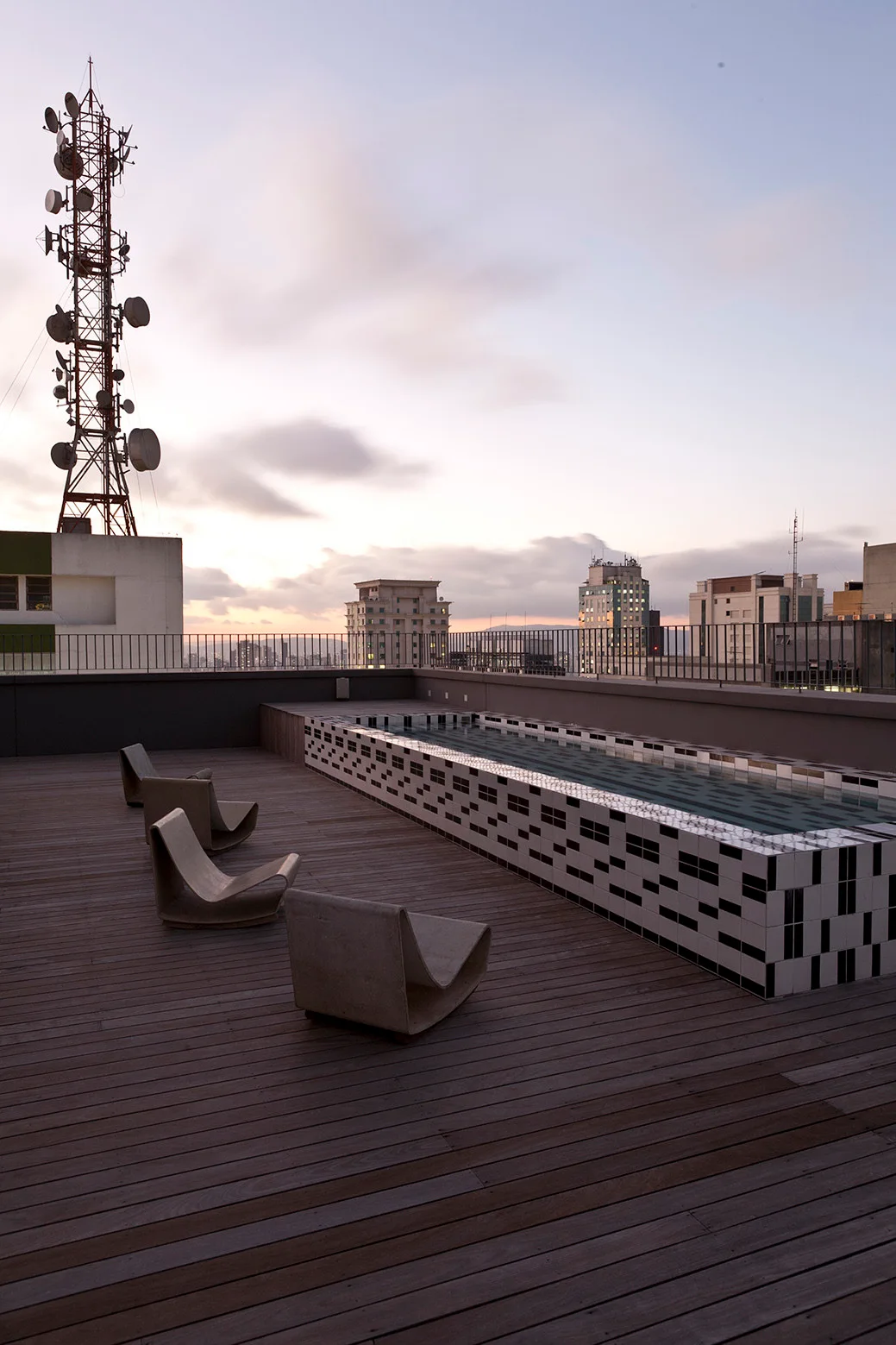 A rooftop wooden deck features low curved concrete lounge chairs beside a raised swimming pool clad in geometric black and white tiles.