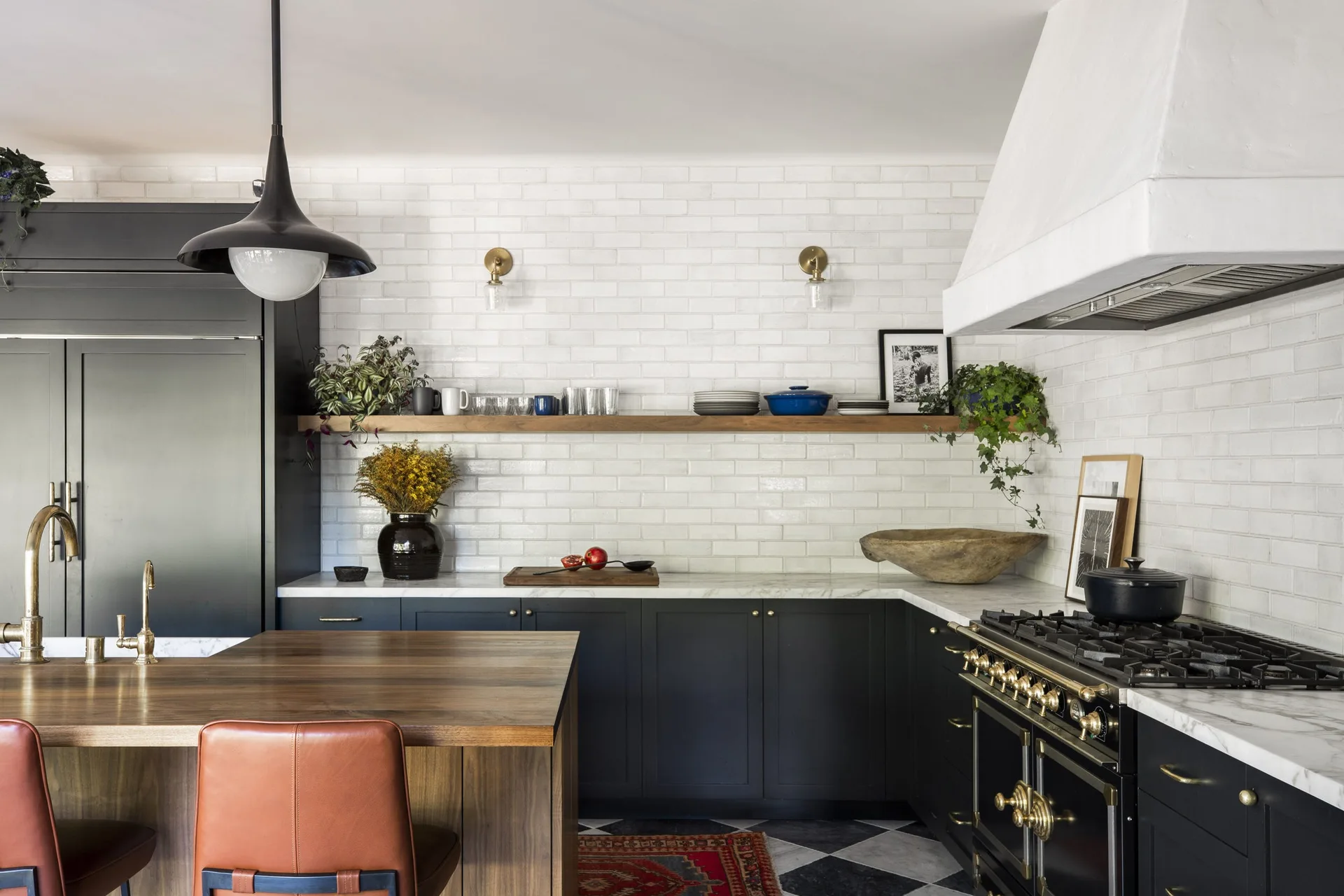 A kitchen featuring dark lower cabinetry, a white tile backsplash, a floating wooden shelf, and a wooden island in the foreground with a leather stool.